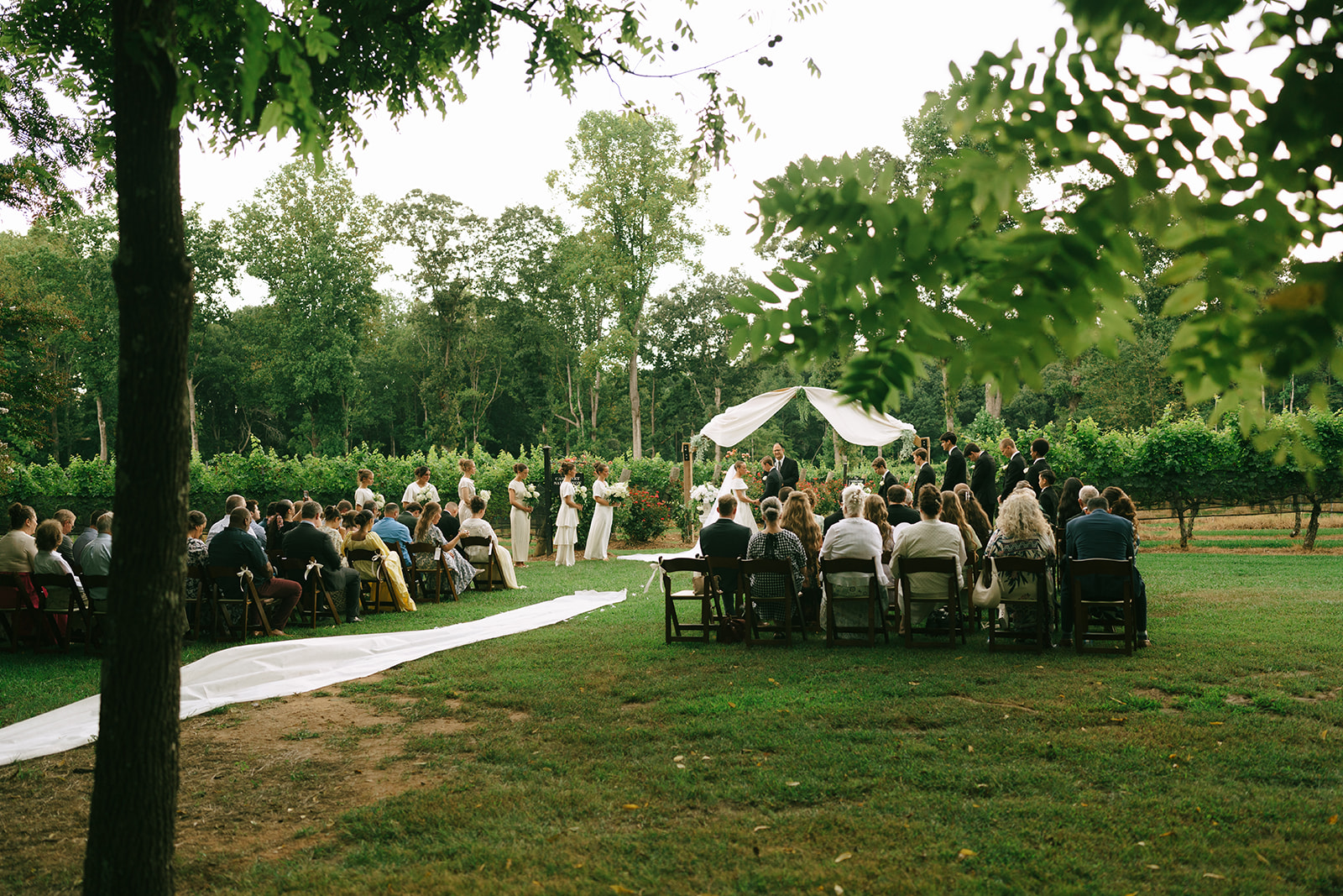 Outdoor wedding ceremony with guests seated on either side of a white aisle, trees and greenery in the background, and a decorated arch at the front.
