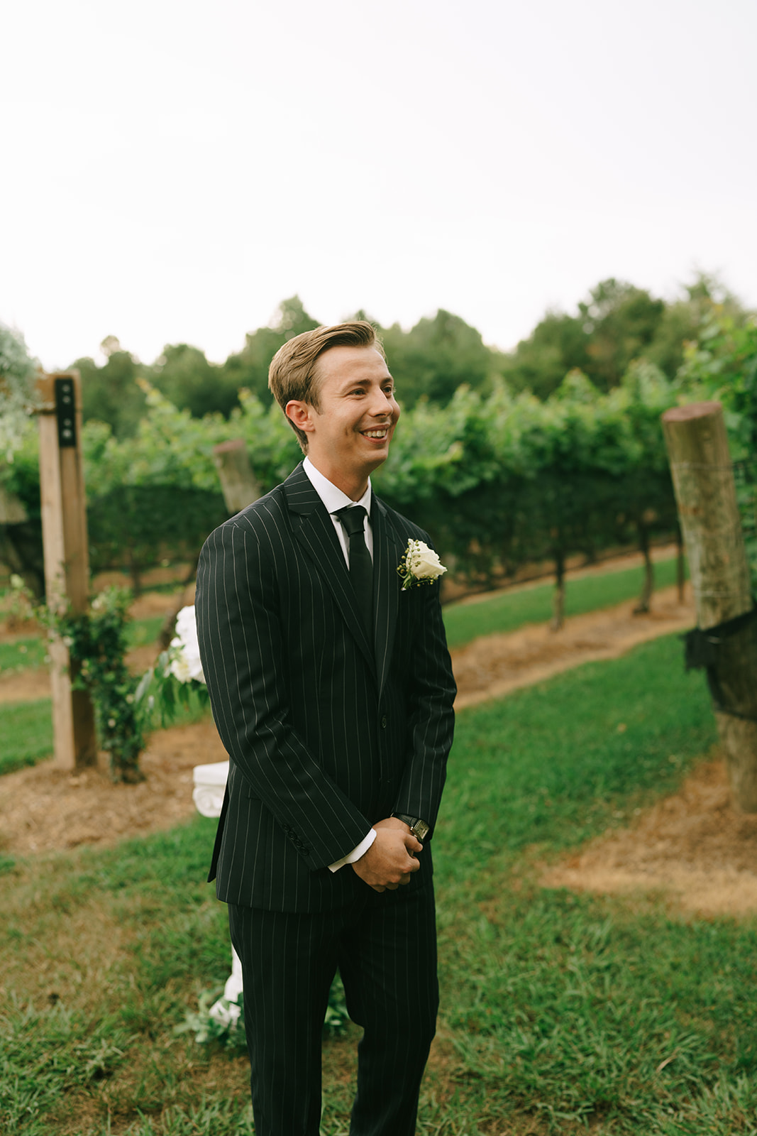 A man in a dark pinstripe suit stands on grass in a vineyard, smiling, with a white boutonniere on his lapel.