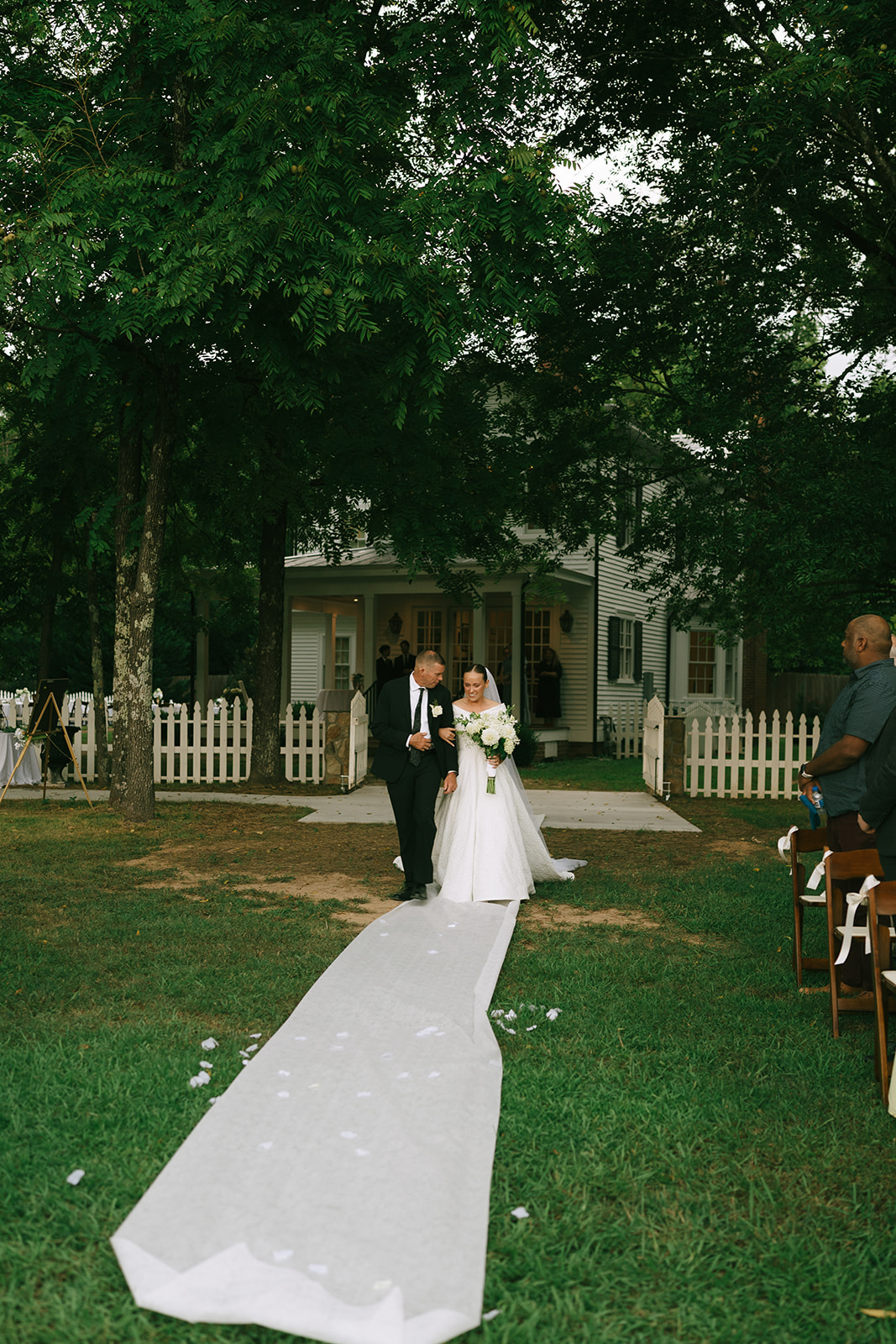 A bride in a white dress walks down an outdoor aisle with an older man, likely her father, towards a white house with a picket fence, surrounded by greenery.