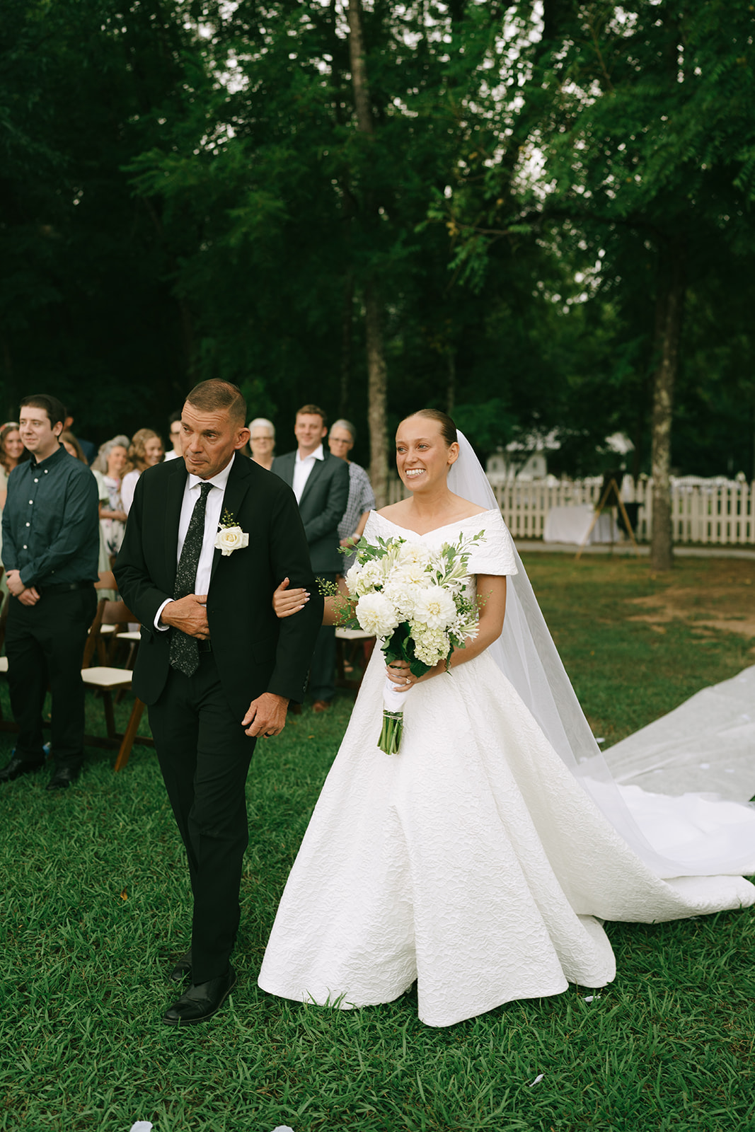 A bride in a white dress walks down an outdoor aisle with an older man, likely her father, towards a white house with a picket fence, surrounded by greenery.