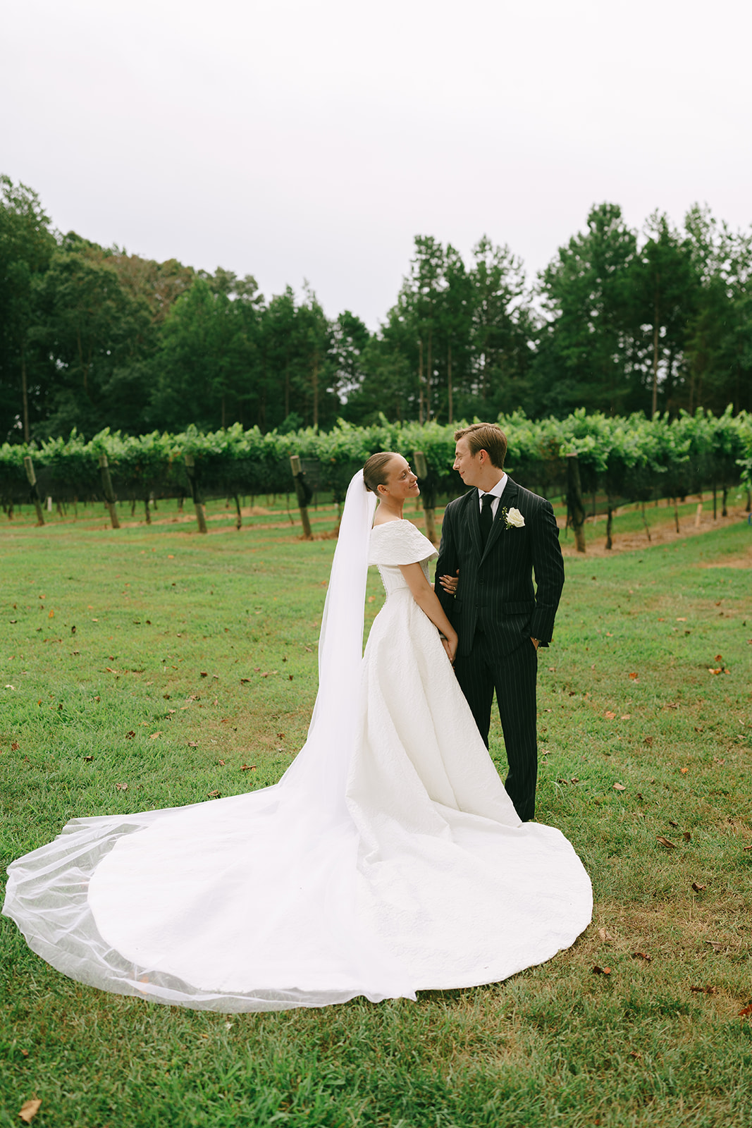 A bride in a white gown and veil stands next to a groom in a dark pinstriped suit, posing outdoors in a green vineyard setting.