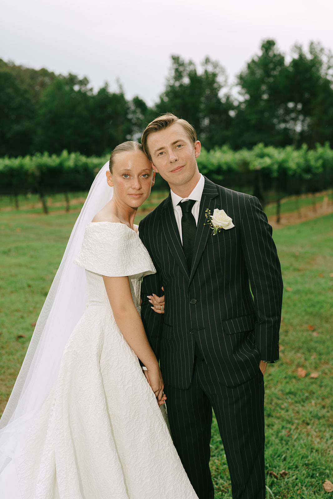 A bride in a white gown and veil stands next to a groom in a dark pinstriped suit, posing outdoors in a green vineyard setting.
