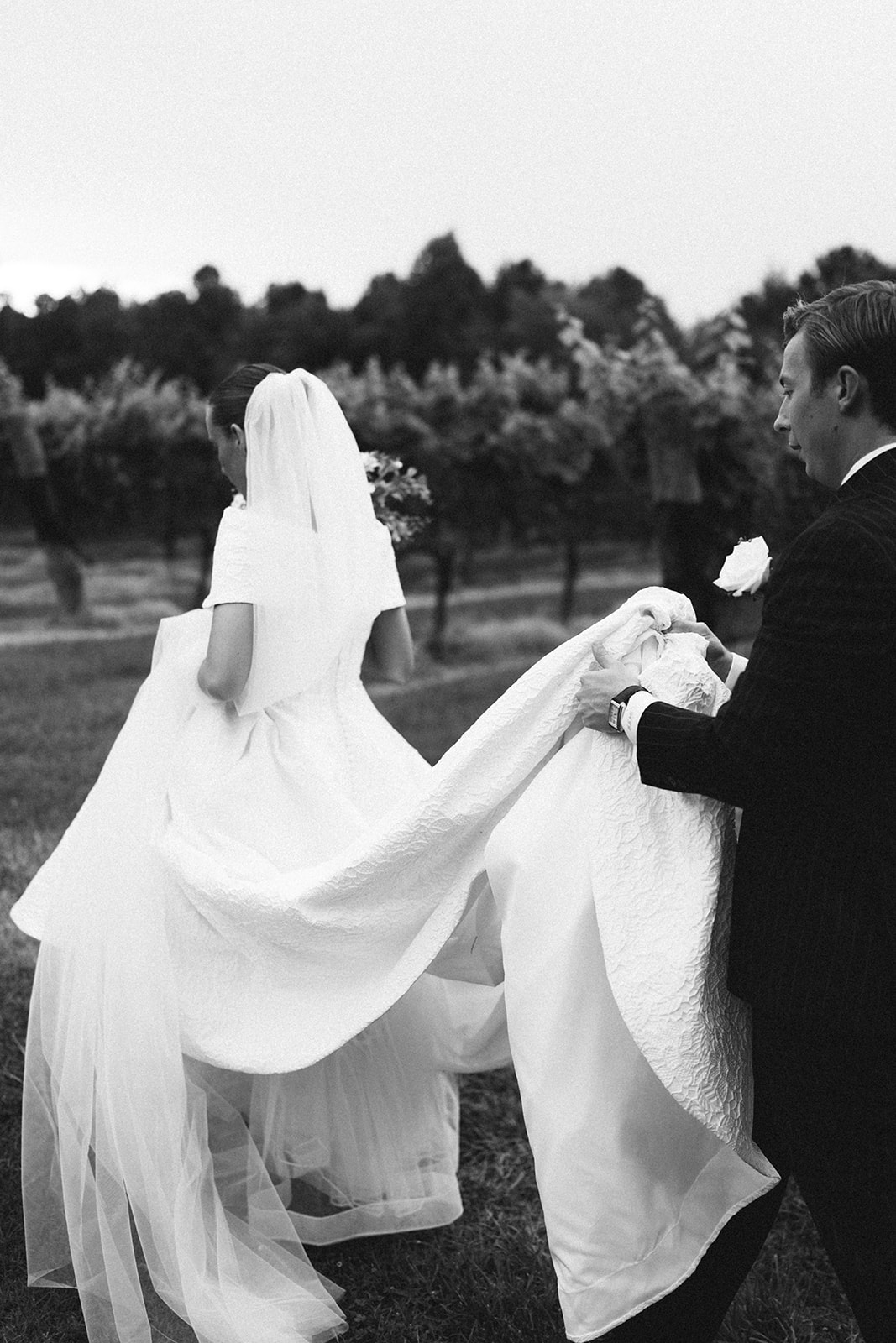 A bride in a white gown and veil stands next to a groom in a dark pinstriped suit, posing outdoors in a green vineyard setting.