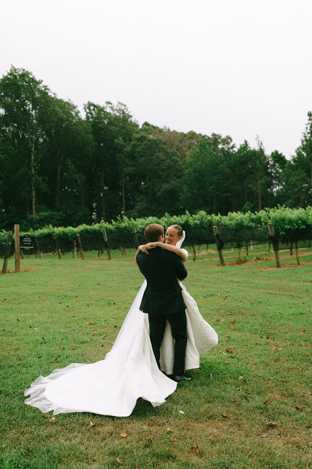 A bride in a white gown and veil stands next to a groom in a dark pinstriped suit, posing outdoors in a green vineyard setting.