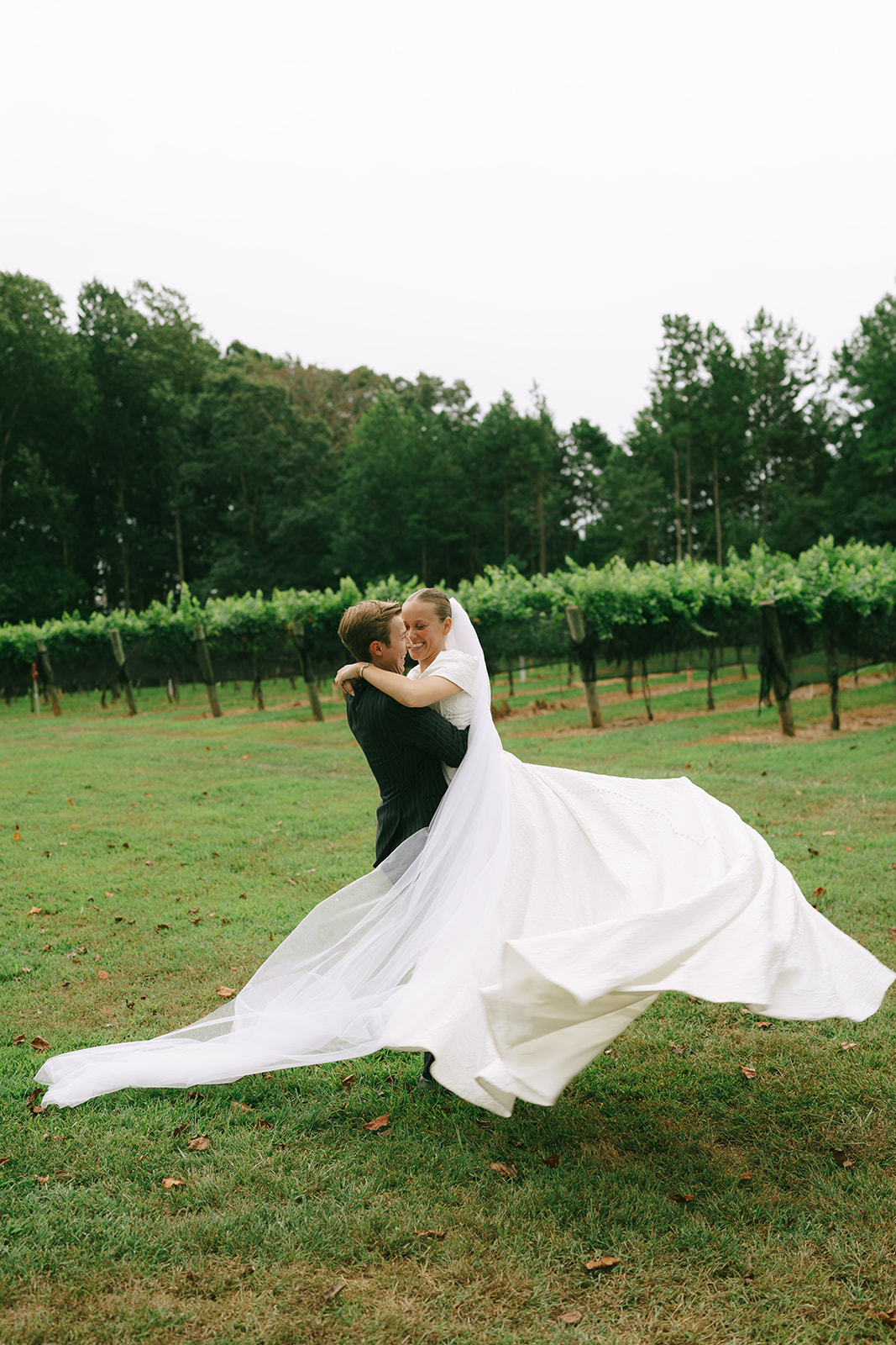 A bride in a white gown and veil stands next to a groom in a dark pinstriped suit, posing outdoors in a green vineyard setting.