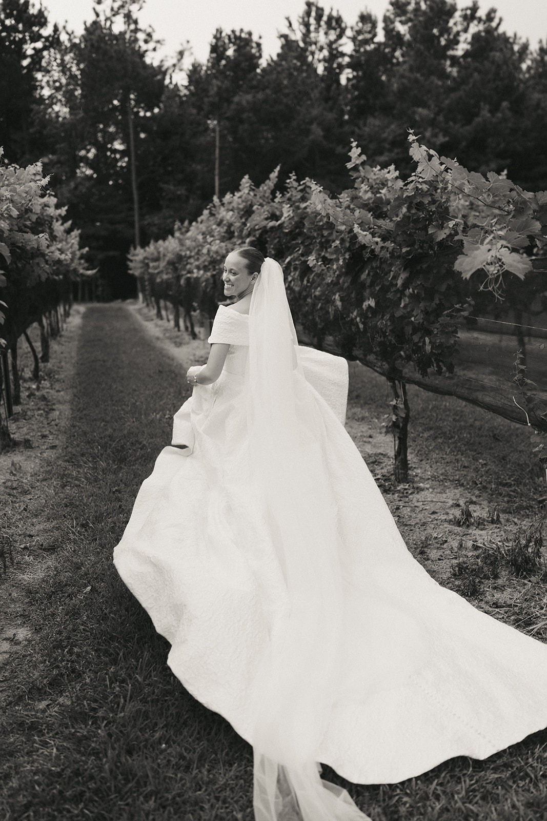 A bride in a white gown and veil stands next to a groom in a dark pinstriped suit, posing outdoors in a green vineyard setting.