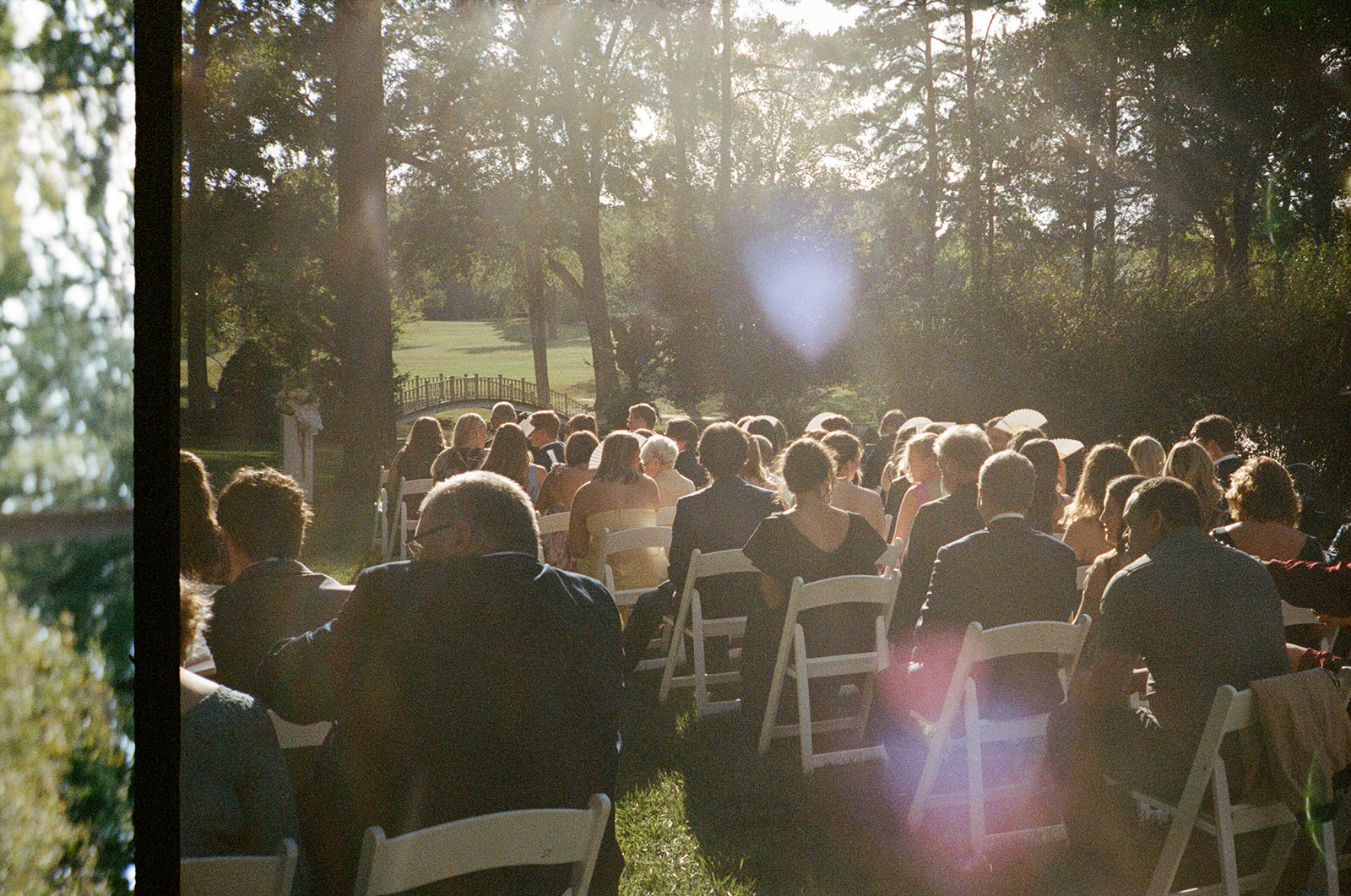 A group of people sits on white chairs outdoors at a sunlit event, possibly a wedding ceremony, with trees and greenery in the background.