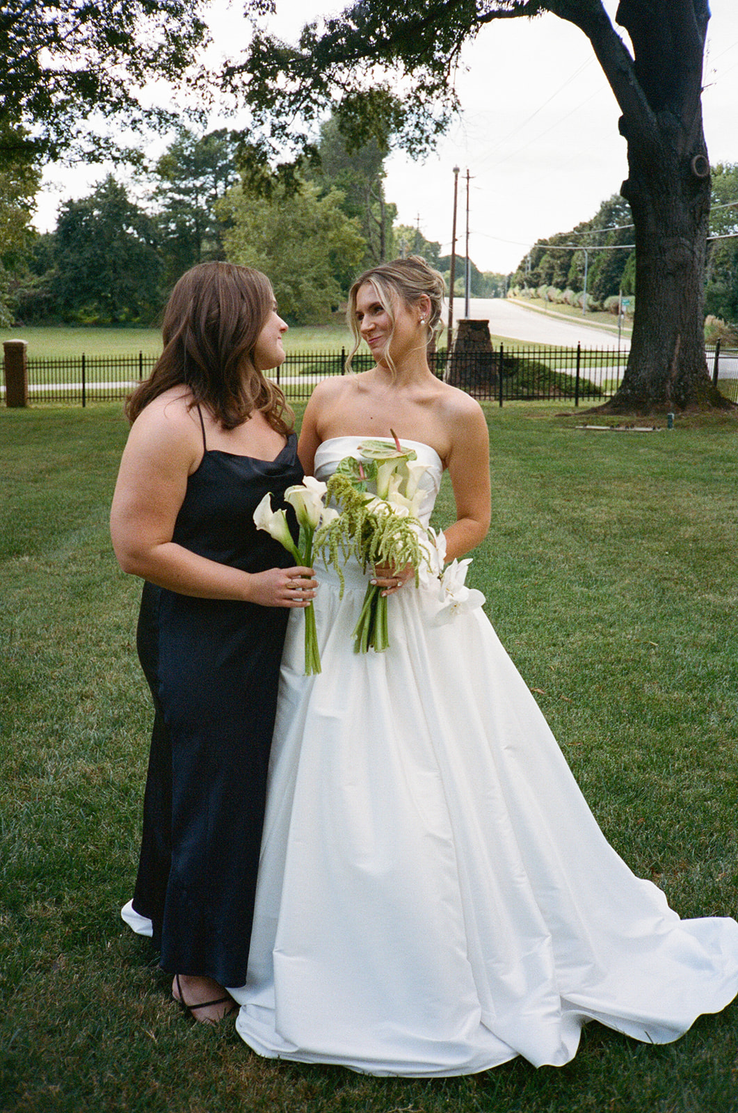 A bride in a white strapless gown stands outdoors among bridesmaids wearing black dresses, holding small bouquets, with trees in the background for a Charleston wedding