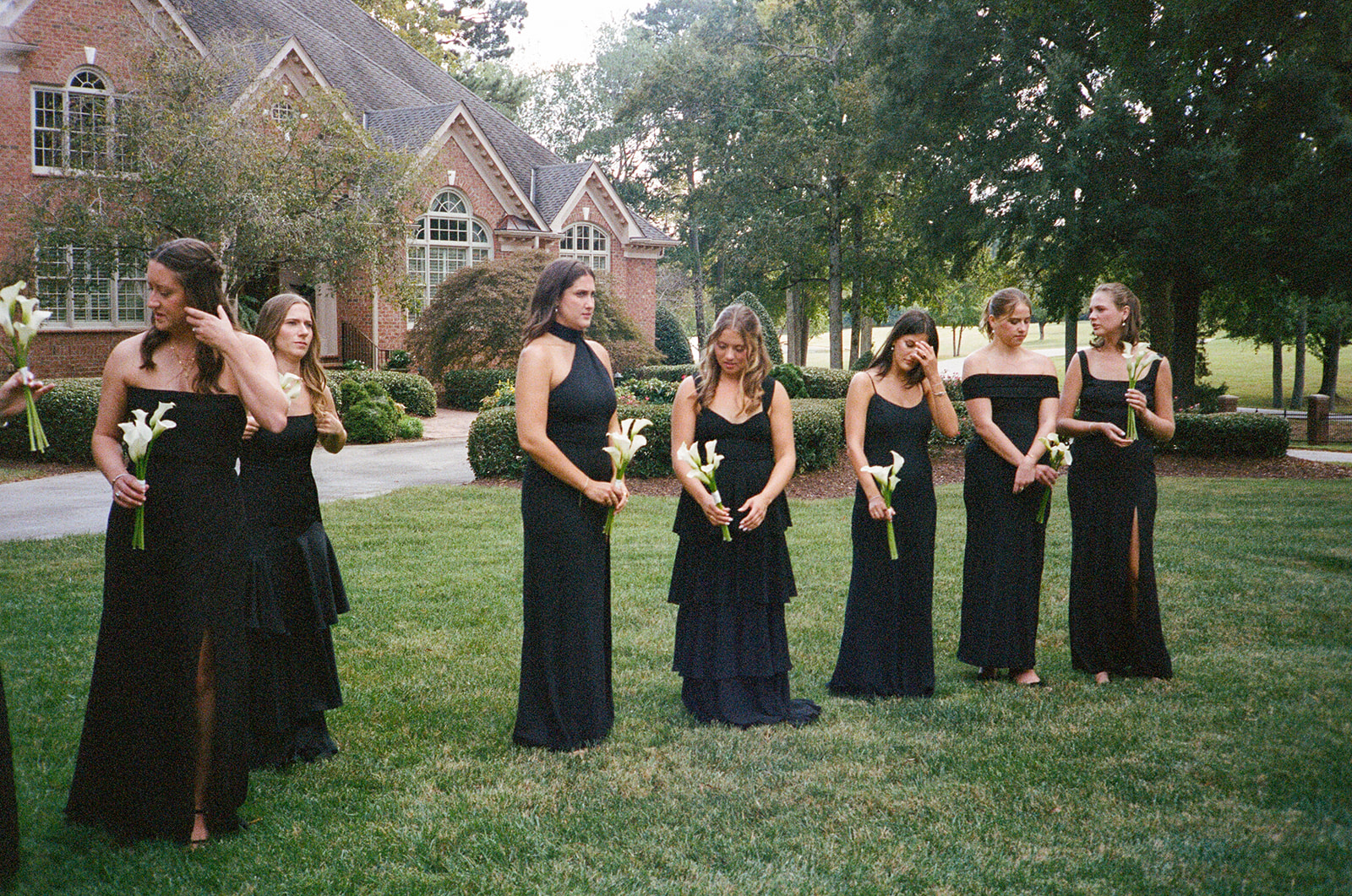 Seven women in black dresses holding white flowers stand on grass in front of a brick house, likely participating in a wedding or formal event.