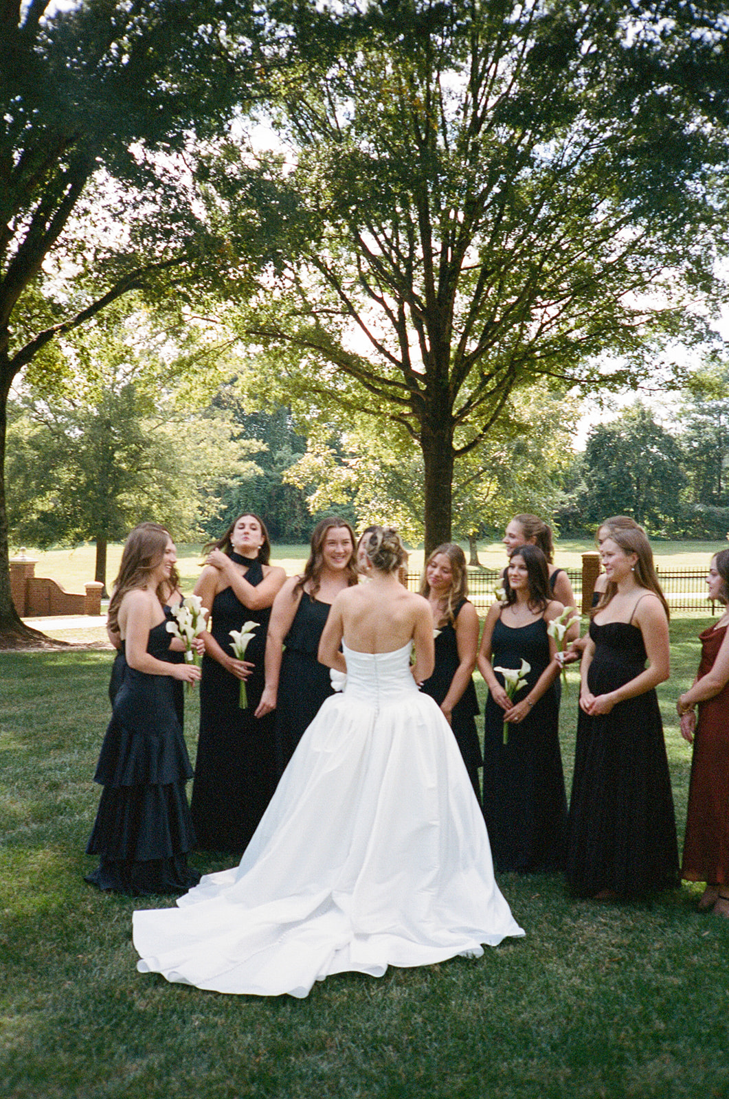 A bride in a white strapless gown stands outdoors among bridesmaids wearing black dresses, holding small bouquets, with trees in the background for a Charleston wedding 