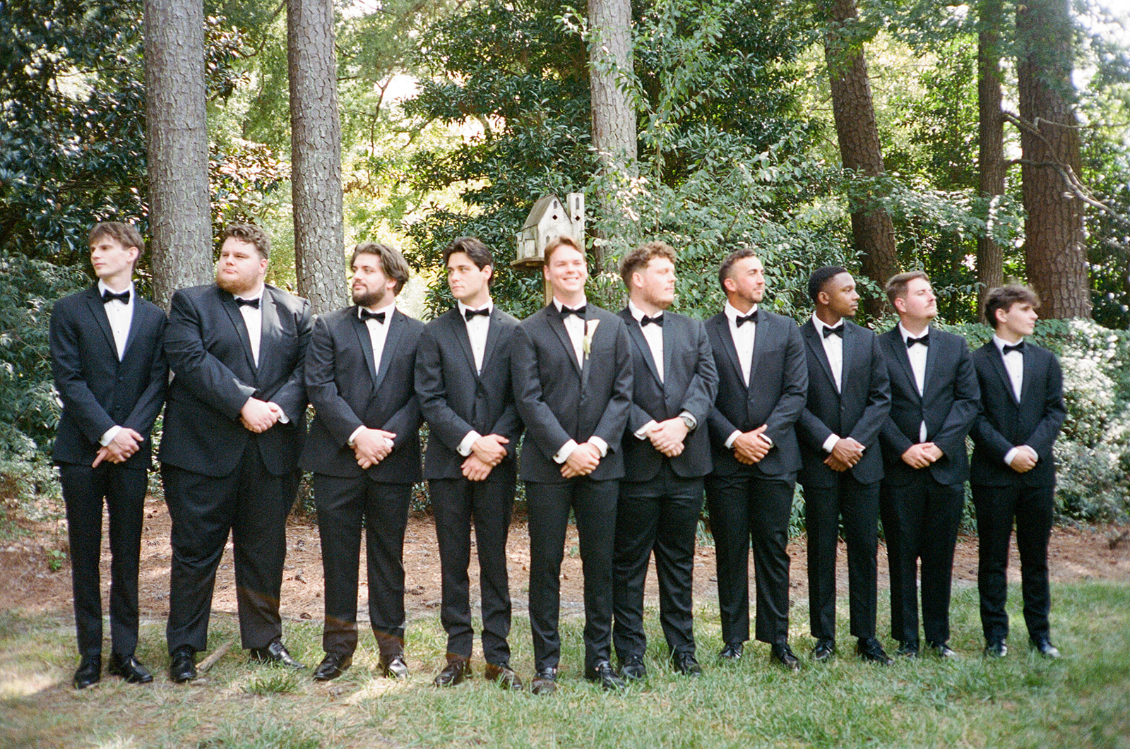 Ten men in black tuxedos stand in a line outdoors, posing for a formal group photo with trees and greenery in the background.