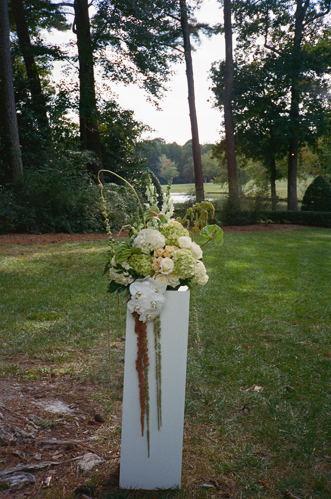 Two tall white flower arrangements stand on pedestals in a grassy outdoor area with trees and a field in the background.