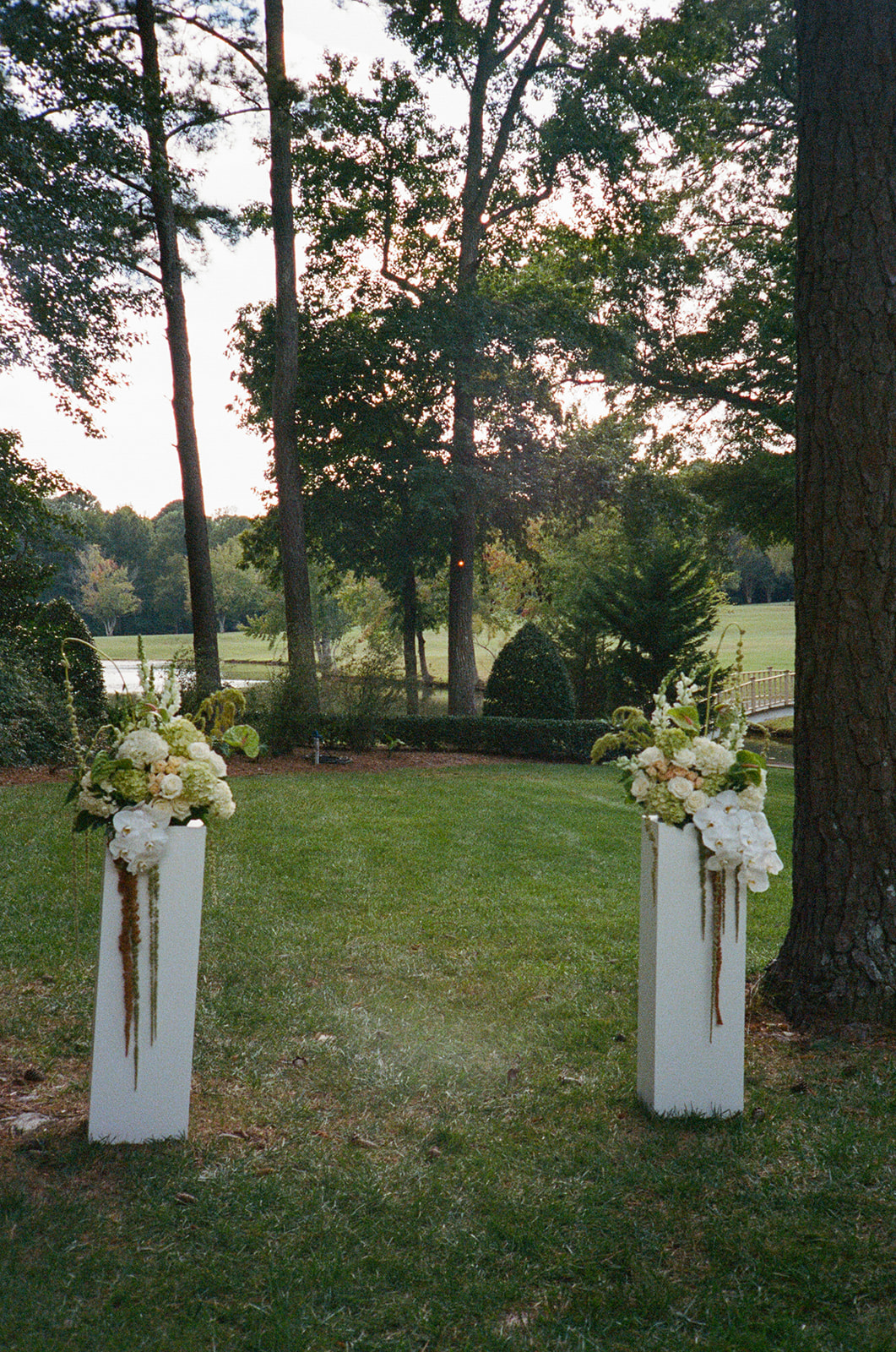 Two tall white flower arrangements stand on pedestals in a grassy outdoor area with trees and a field in the background.