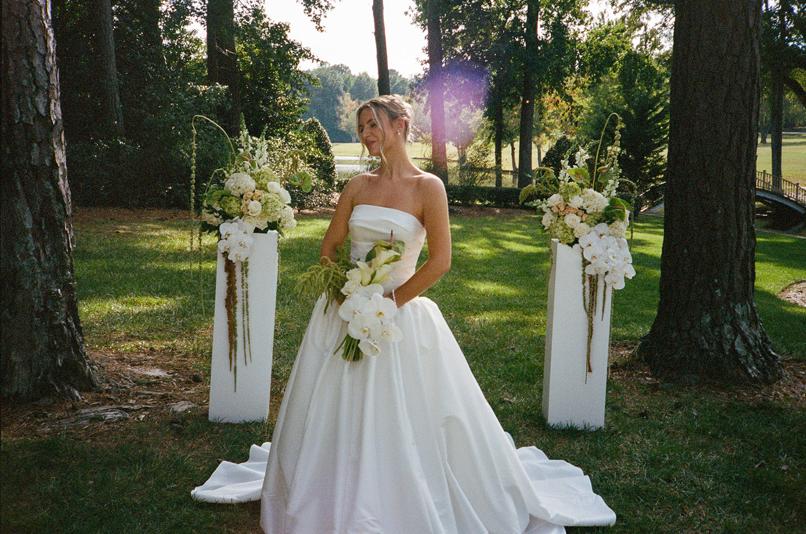 A bride in a white wedding dress stands outdoors on grass, holding a bouquet, with two floral arrangements on white pedestals behind her for a Charleston wedding