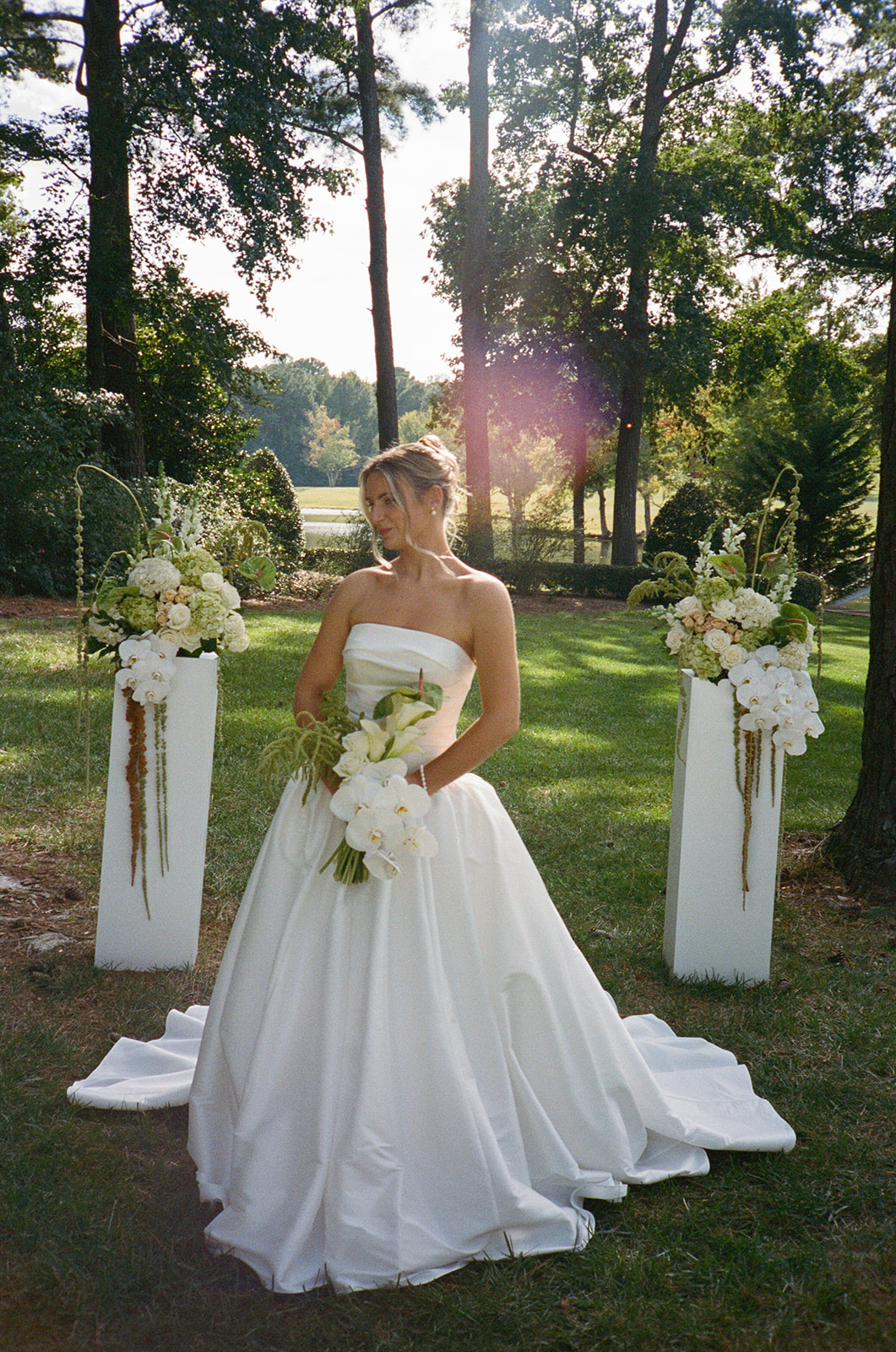 A woman in a strapless white dress stands outdoors on grass, holding a bouquet and smiling, in a black and white photograph.