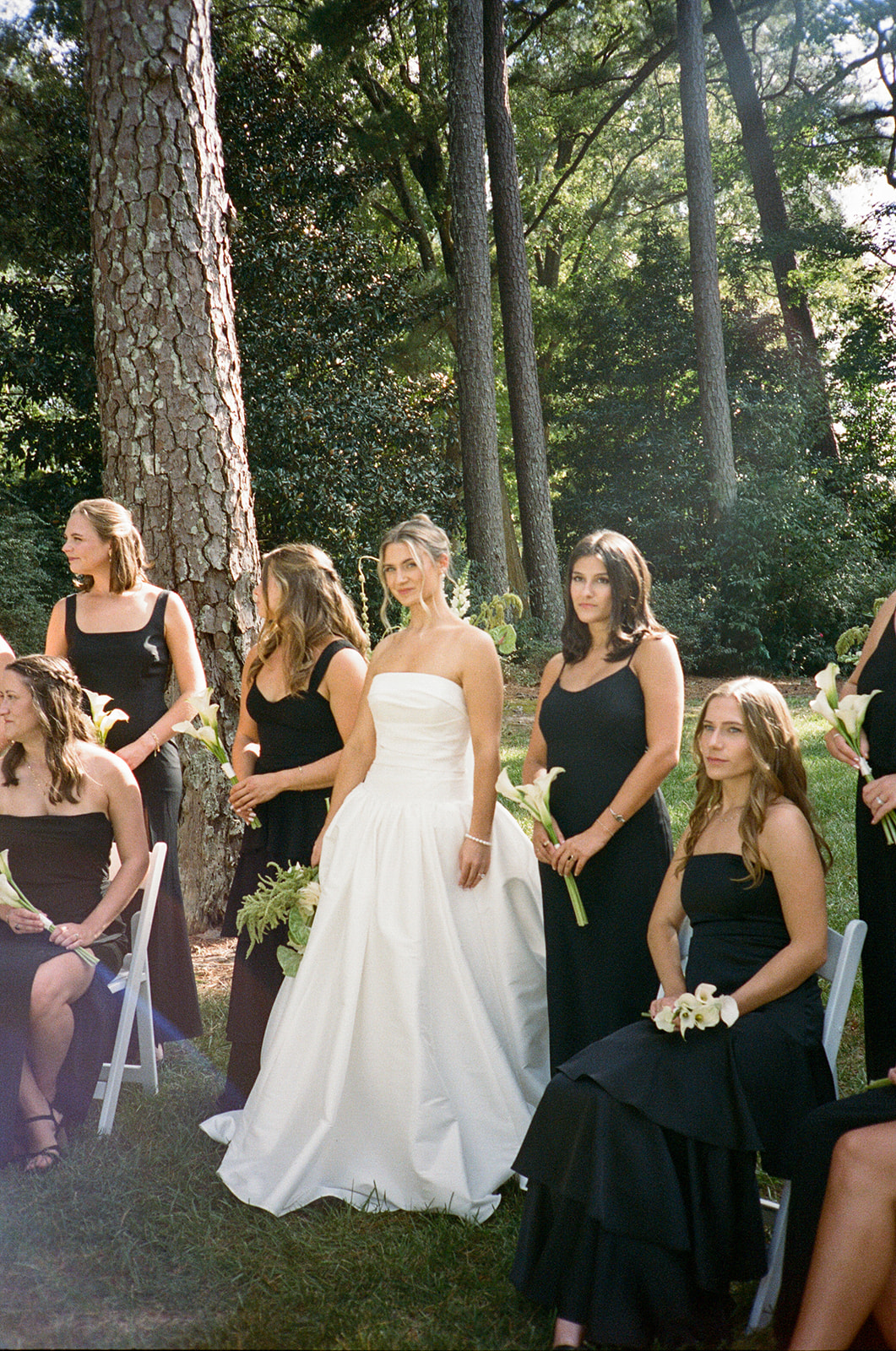 A bride in a white strapless gown stands outdoors among bridesmaids wearing black dresses, holding small bouquets, with trees in the background for a Charleston wedding 