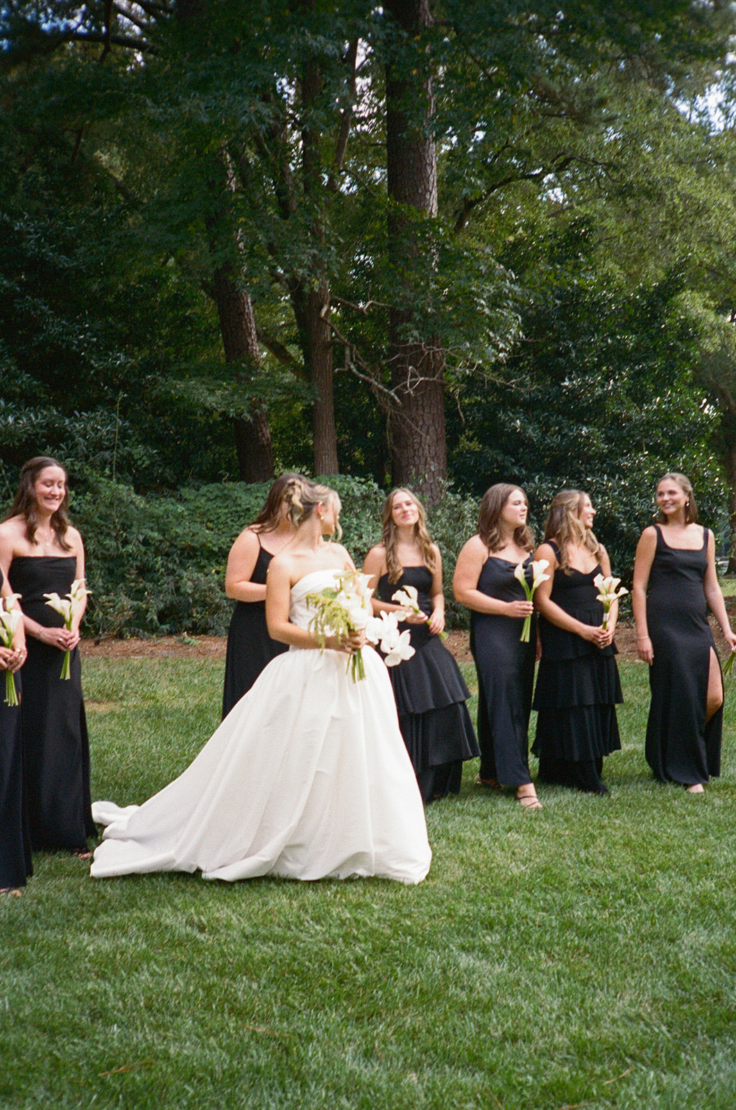 A bride in a white strapless gown stands outdoors among bridesmaids wearing black dresses, holding small bouquets, with trees in the background for a Charleston wedding