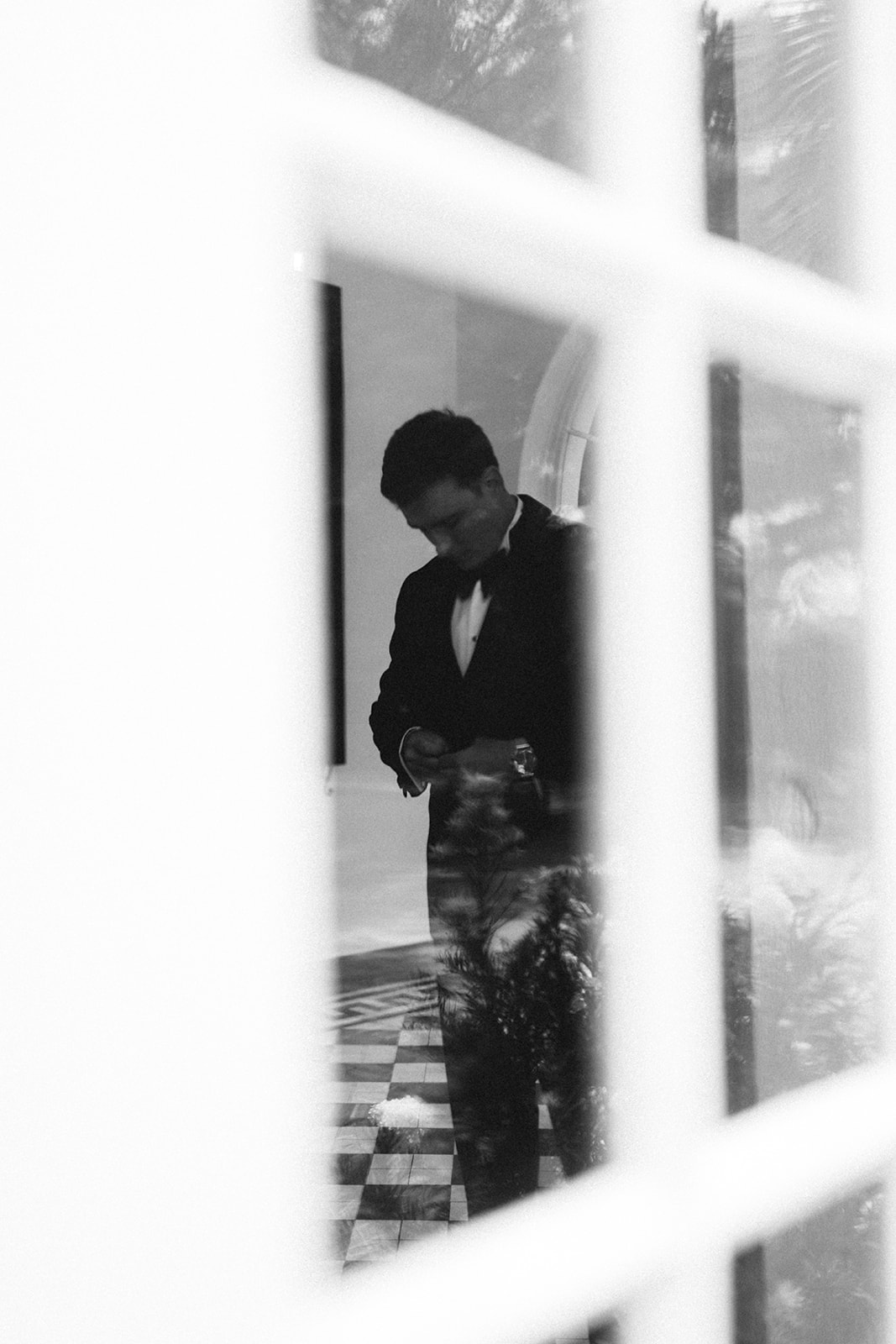 Black and white photo of a man in a suit, seen through a window with grid panes, standing indoors on a checkered floor.