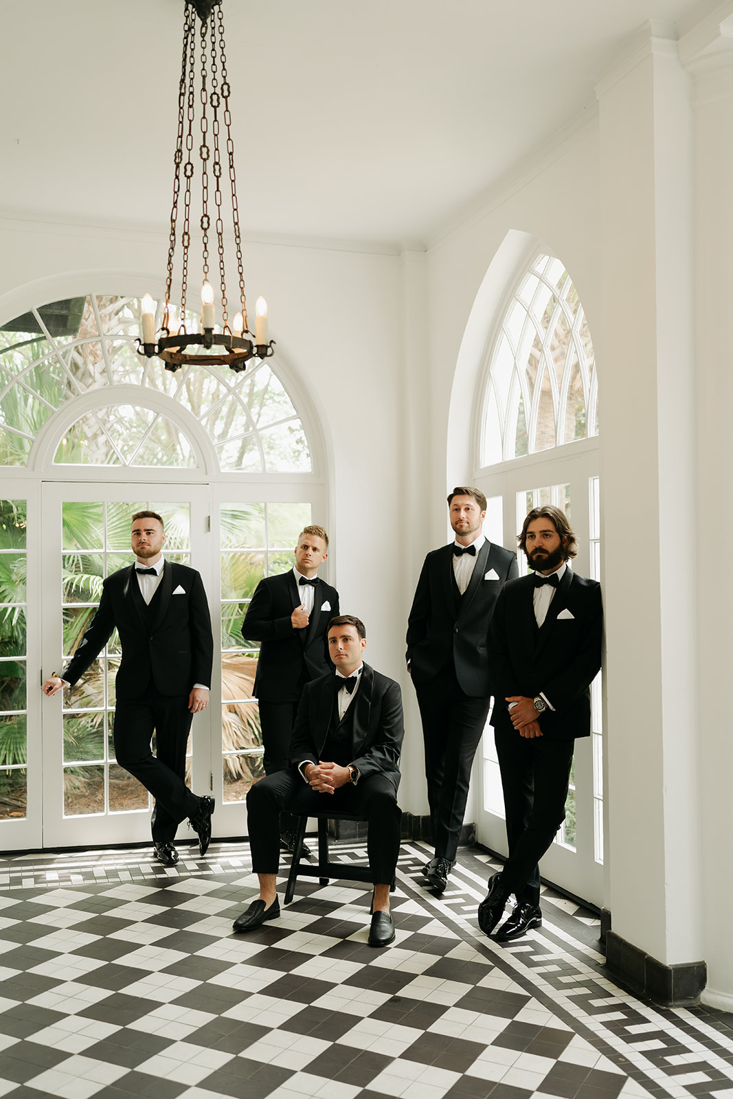 Five men in black tuxedos pose in a bright room with checkered floor and large windows; one is seated while the others stand around him.