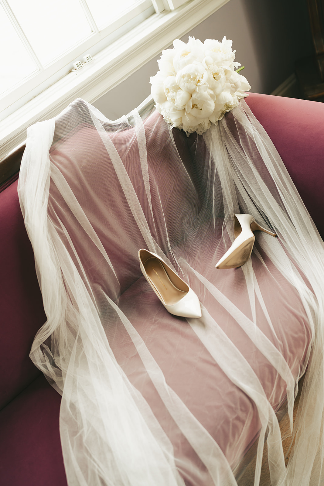 A bridal veil draped over a mauve chair with a bouquet of white flowers and a pair of white high-heeled shoes placed on the seat.