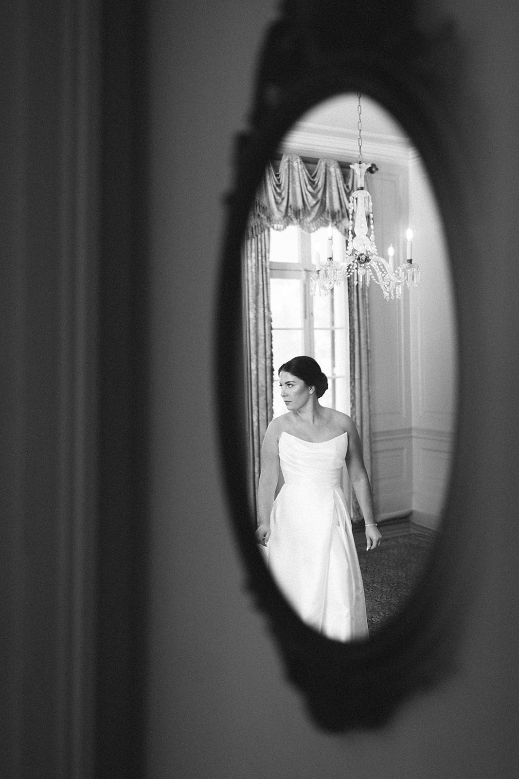 Black and white photo showing a woman in a white dress reflected in an oval mirror, standing in a room with large windows and a chandelier.