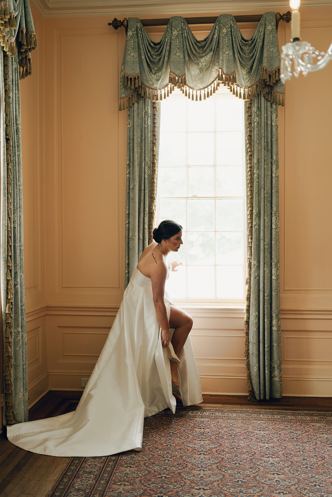 A bride in a white dress sits on a chair in front of a fireplace, holding a bouquet of flowers in an elegant, vintage-style room at a wedding venue in charleston