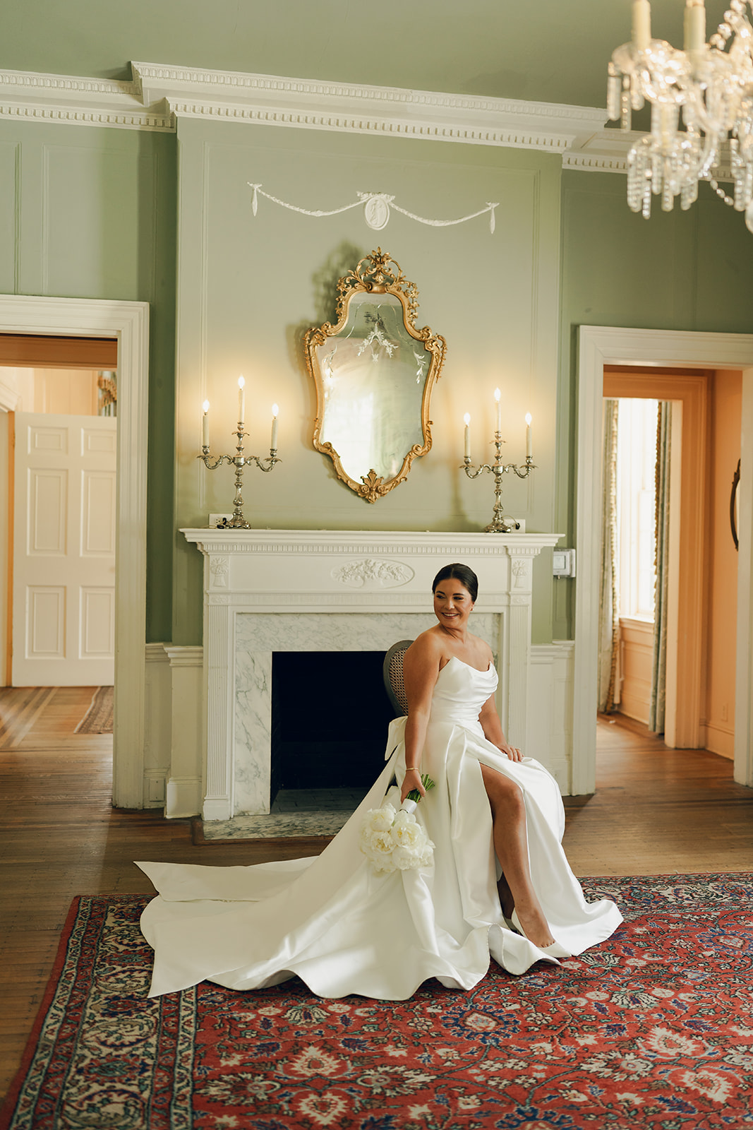 A bride in a white dress sits on a chair in front of a fireplace, holding a bouquet of flowers in an elegant, vintage-style room at a wedding venue in charleston