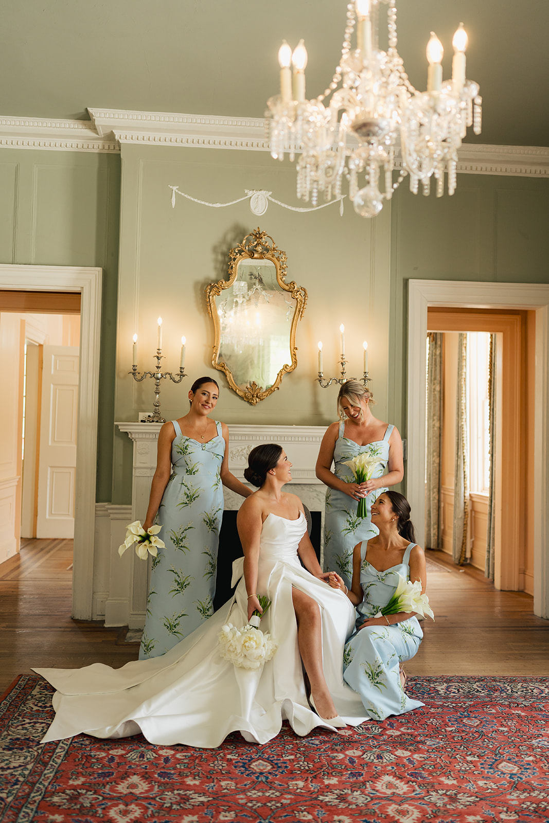 A bride in a white gown sits on a chair, surrounded by three bridesmaids in matching blue dresses, holding white flowers in an elegant room with a chandelier.