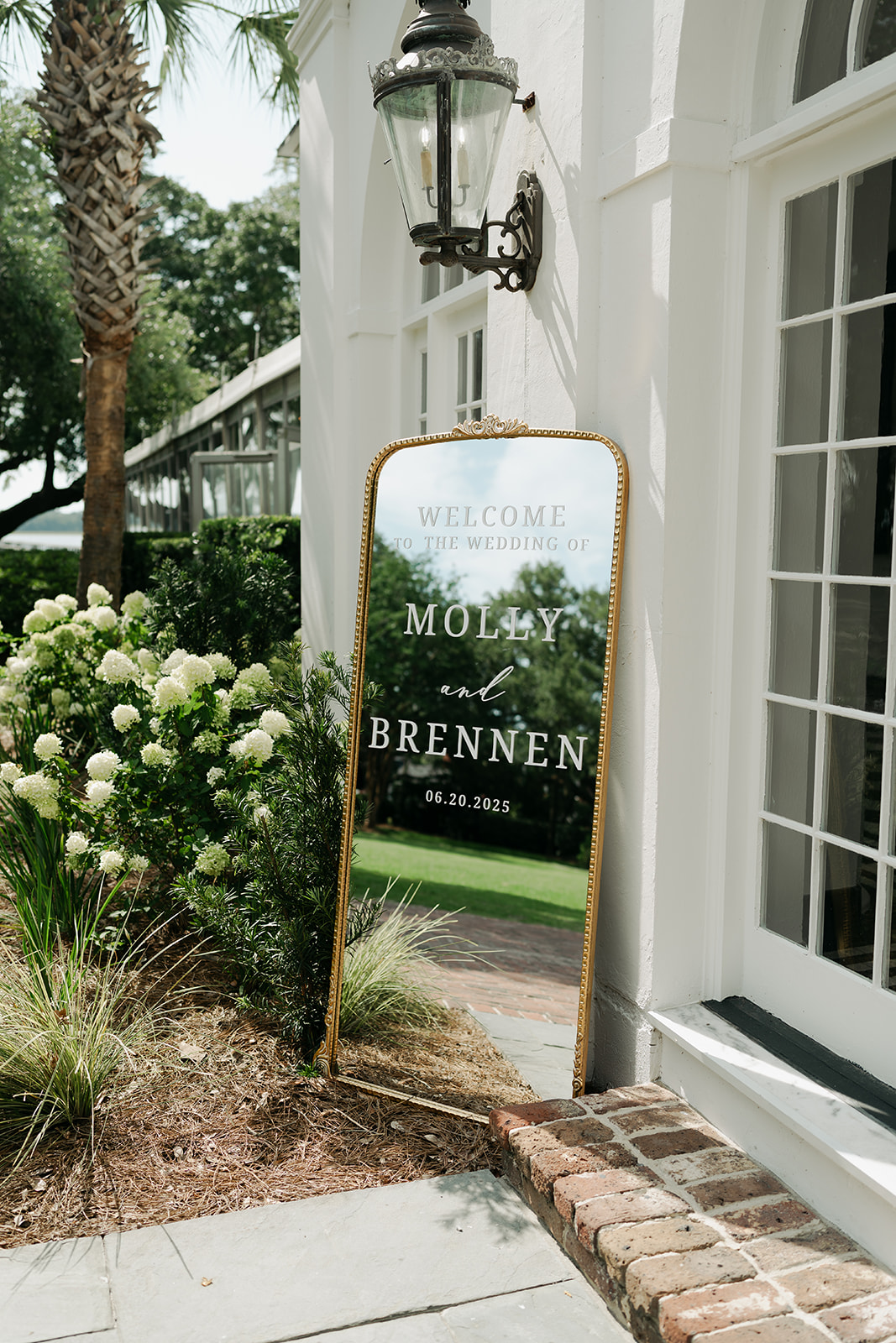 A wedding welcome sign for Molly and Brennen, dated 06.24.2023, stands on a sidewalk by a white building with plants and a lantern nearby.