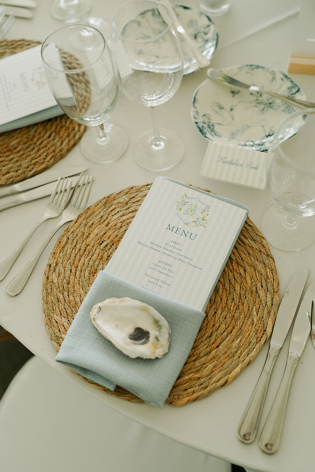 A place setting with a woven placemat, menu card, folded napkin, oyster shell, cutlery, empty glasses, and glass dish on a white tablecloth.