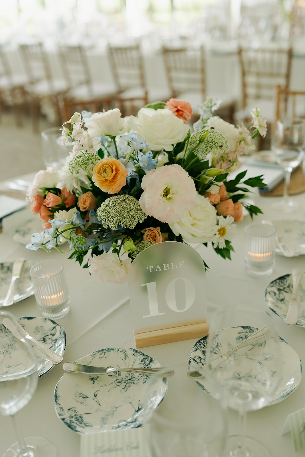 A floral centerpiece with white, peach, and light blue flowers sits on a round table set with plates, glassware, and a table number 10 sign at an event.