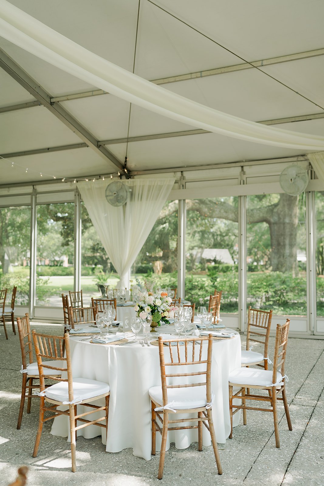 Long banquet table with white tablecloth, elegant table settings, tall floral centerpieces, and wooden chairs under a white tent with draped fabric and string lights for a wedding venue in charleston