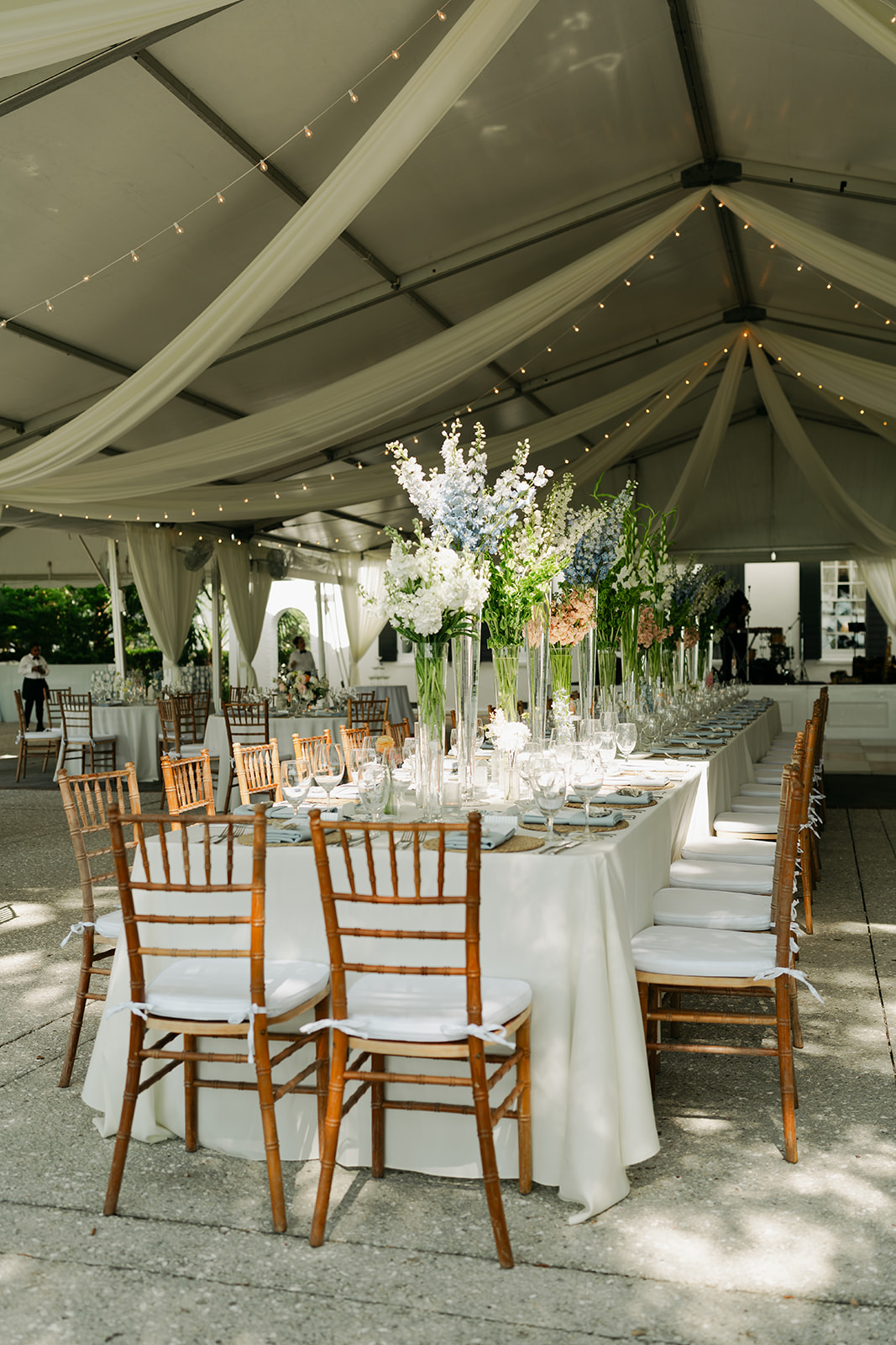 Long banquet table with white tablecloth, elegant table settings, tall floral centerpieces, and wooden chairs under a white tent with draped fabric and string lights for a wedding venue in charleston