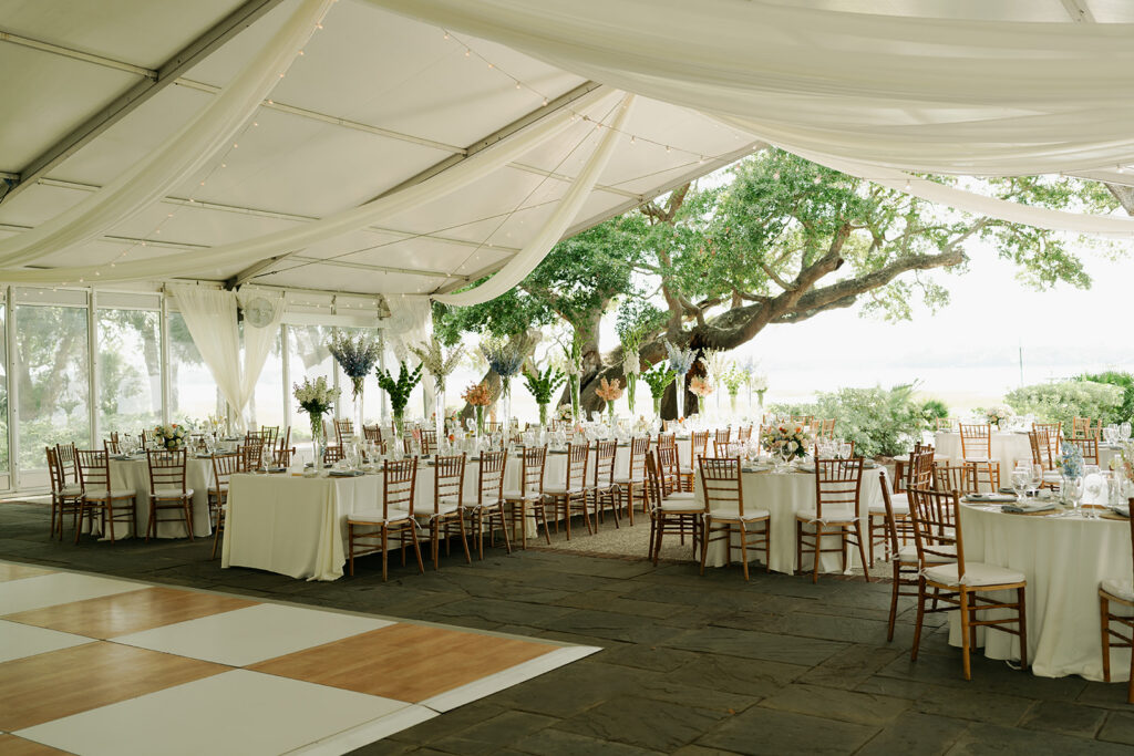 An outdoor event tent with wooden chairs and round tables set for a formal gathering, featuring white linens, floral centerpieces, and a checkered dance floor.
