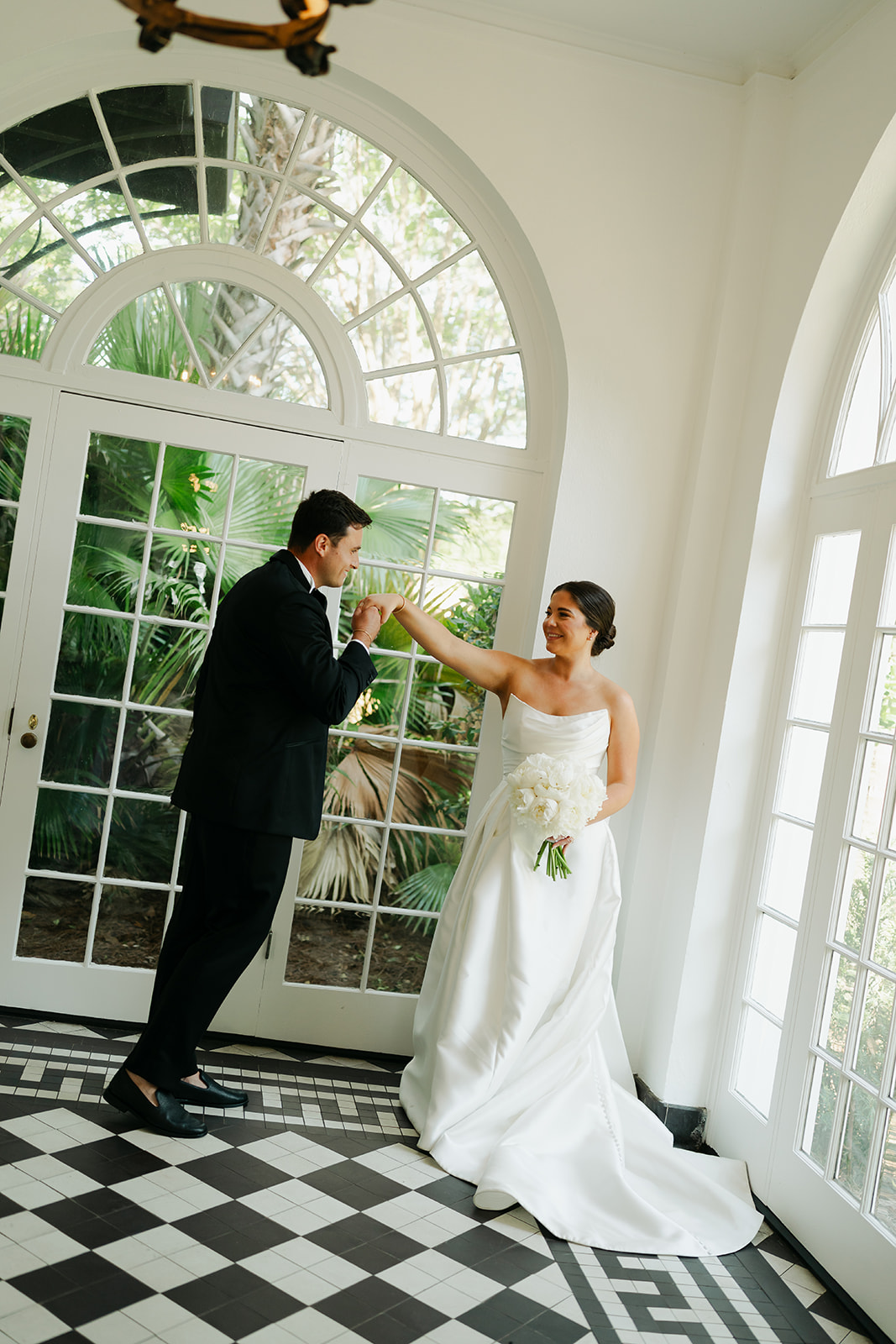 A bride in a white dress and a groom in a black suit stand in a bright room with large windows and black-and-white checkered floor, holding hands and smiling at each other at Wedding Venues in Charleston,