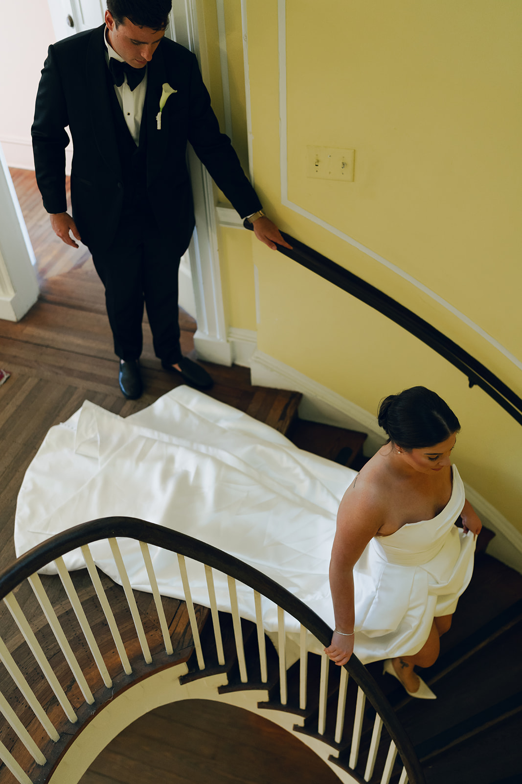 A bride in a white gown ascends a curved wooden staircase, followed by a groom in a black tuxedo at a wedding venue in Charleston
