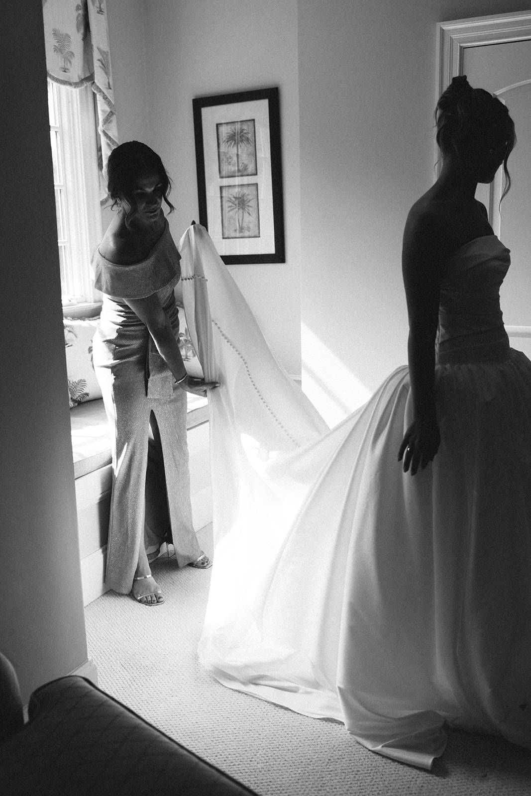 A woman adjusts the train of a bride's wedding dress in a softly lit room.
