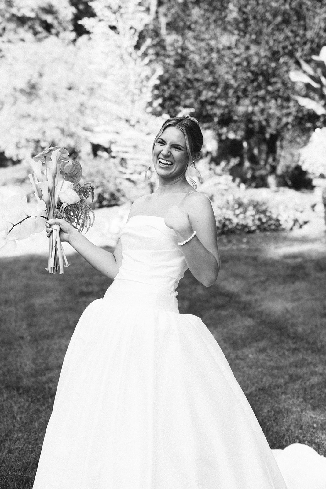 A woman in a strapless white dress stands outdoors on grass, holding a bouquet and smiling, in a black and white photograph.
