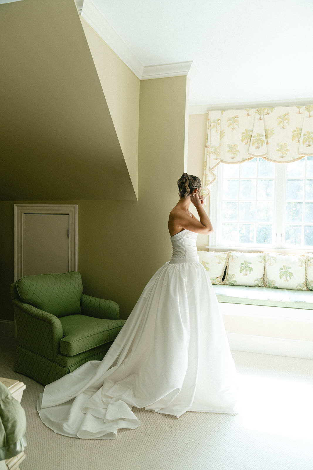 A woman in a strapless white wedding dress stands near a window, adjusting her earring in a softly lit room with green furniture for a Charleston wedding