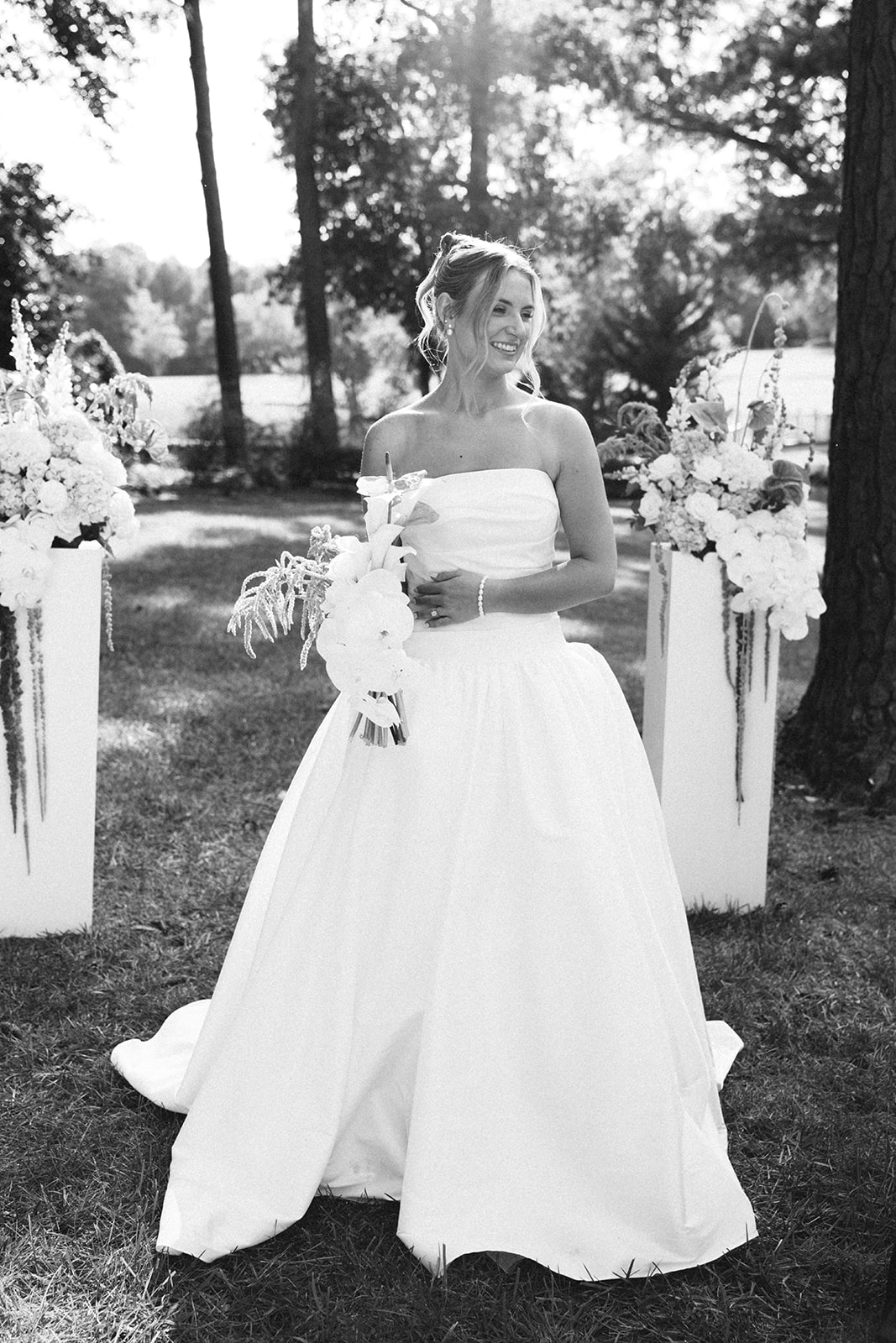 A woman in a strapless white dress stands outdoors on grass, holding a bouquet and smiling, in a black and white photograph.