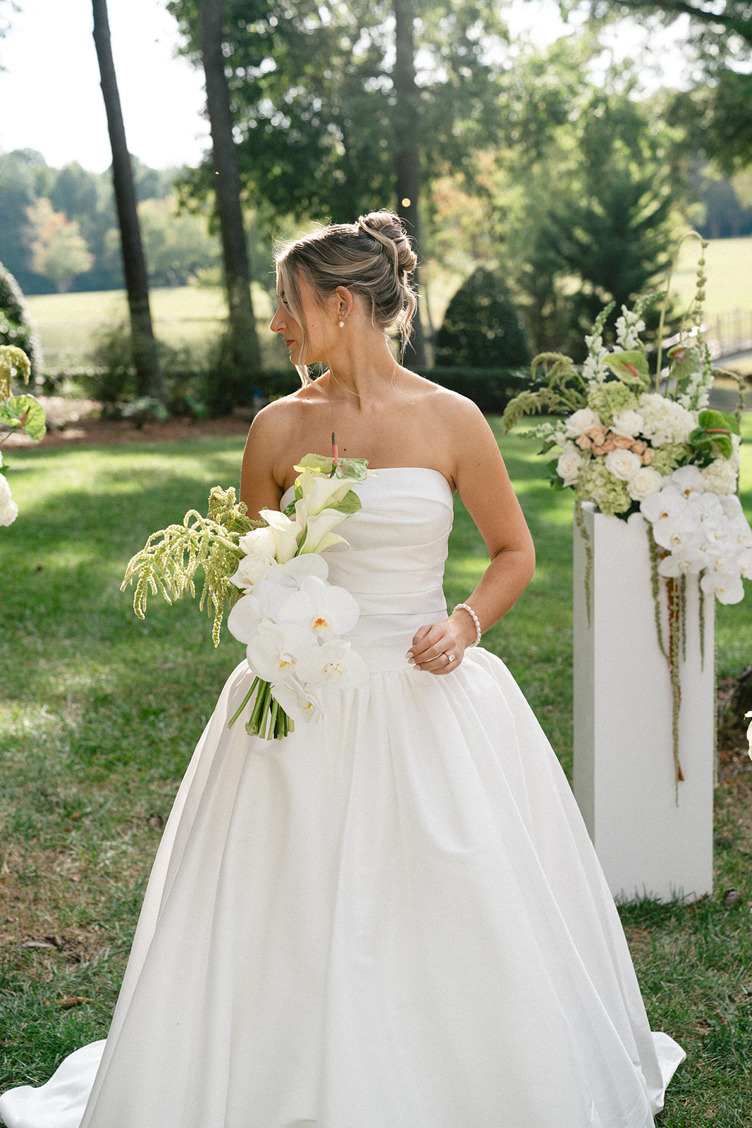 A bride in a white wedding dress stands outdoors on grass, holding a bouquet, with two floral arrangements on white pedestals behind her for a Charleston wedding