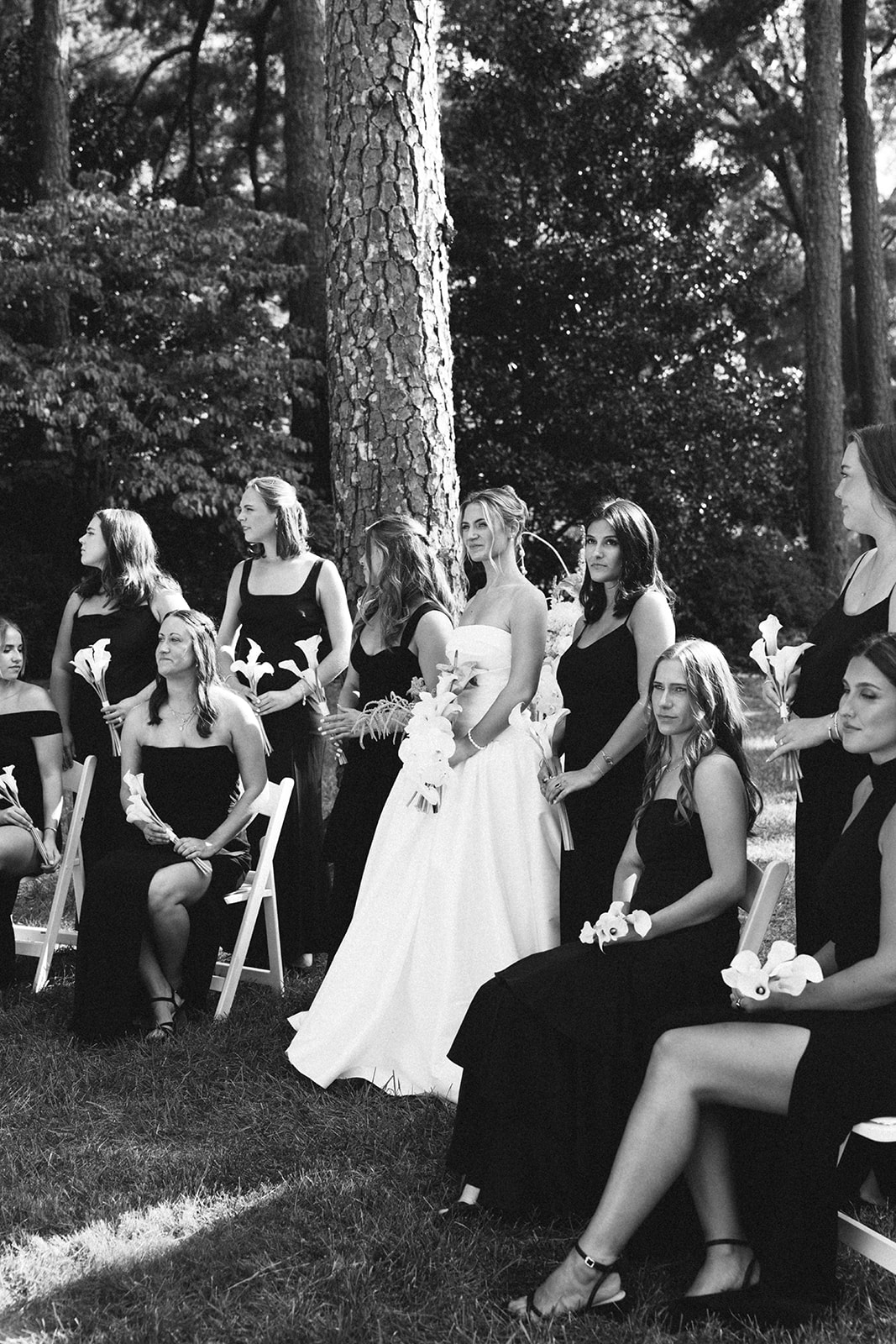 A bride in a white strapless gown stands outdoors among bridesmaids wearing black dresses, holding small bouquets, with trees in the background 