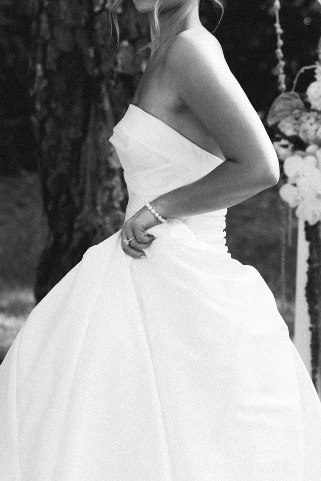 A woman in a strapless white dress stands outdoors on grass, holding a bouquet and smiling, in a black and white photograph.