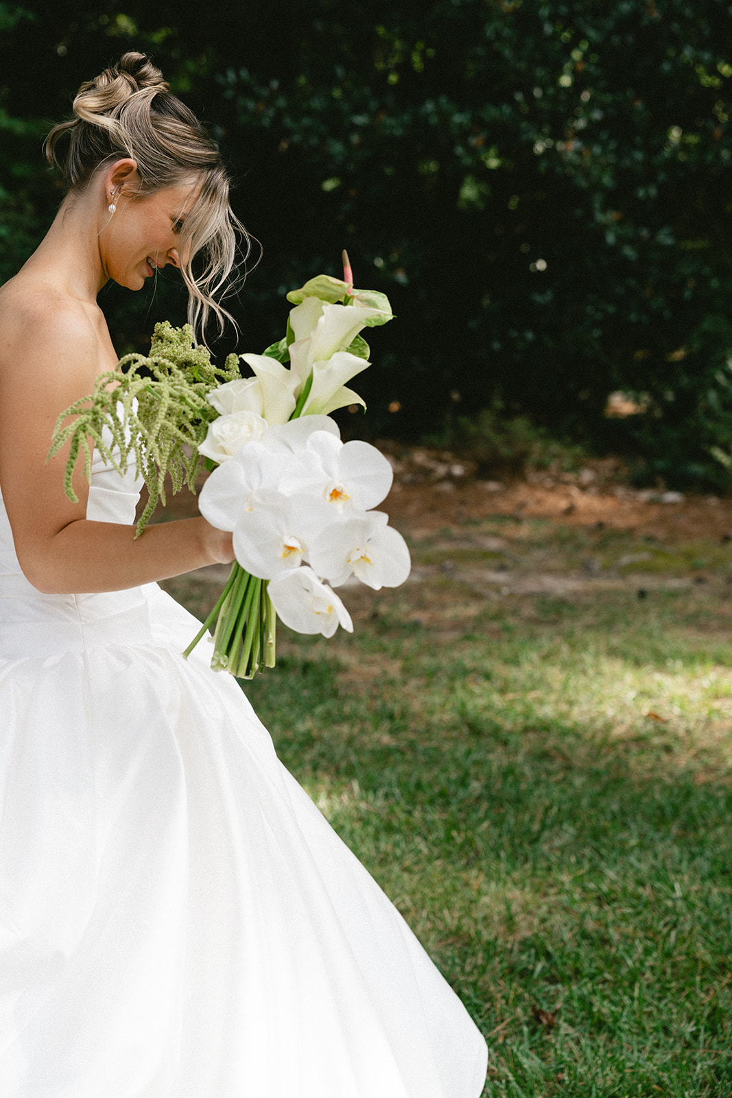 A bride in a white wedding dress stands outdoors on grass, holding a bouquet, with two floral arrangements on white pedestals behind her for a Charleston wedding