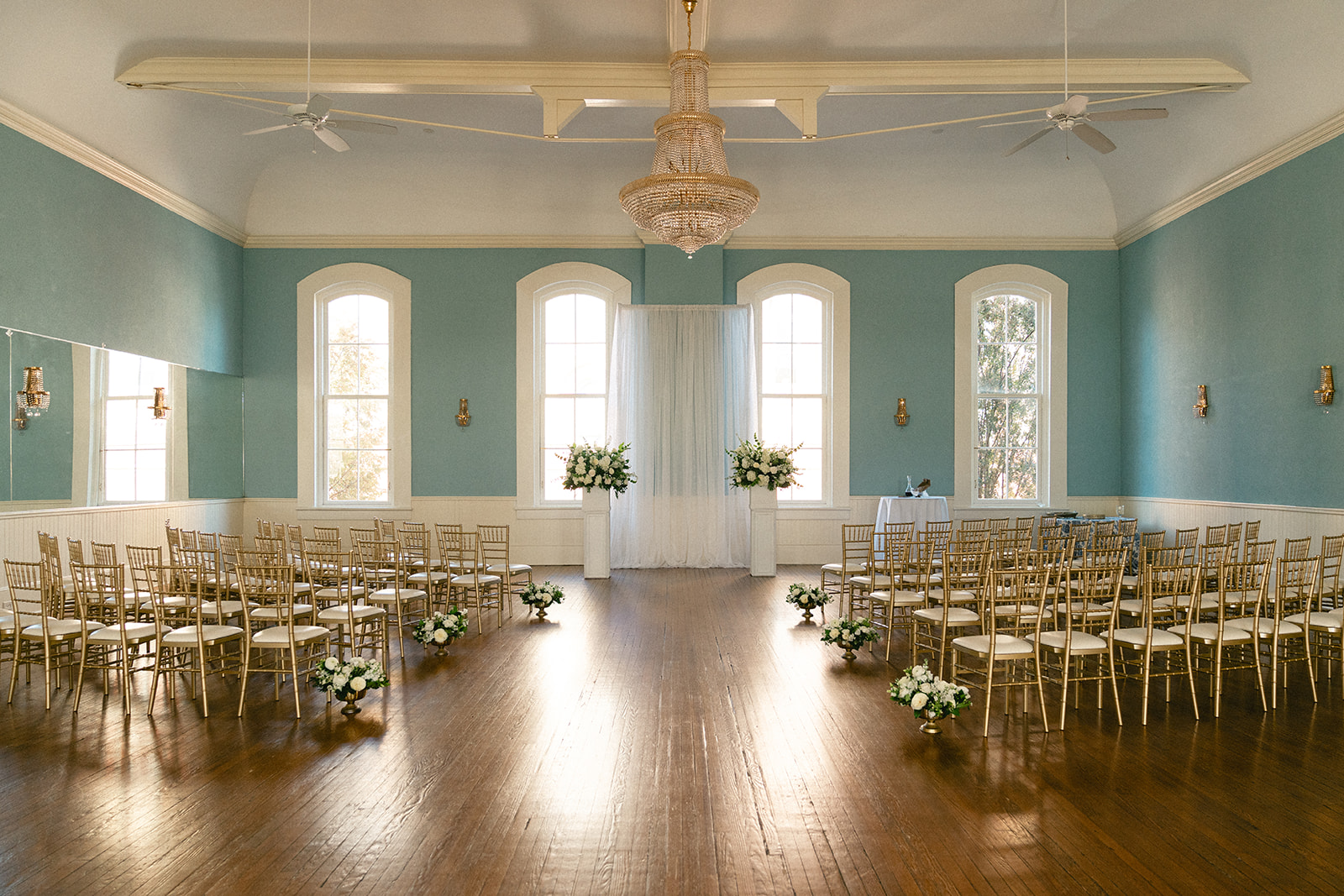 A well-lit room set up for a wedding ceremony, featuring rows of chairs, floral arrangements, and large windows with light blue walls at the Stanly Ballroom a wedding venue in North Carolina