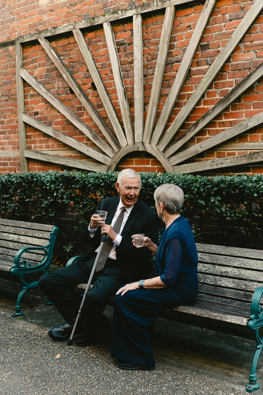 An older man and woman sit on a bench, holding drinks and talking in front of a brick wall with a decorative wooden fan design.