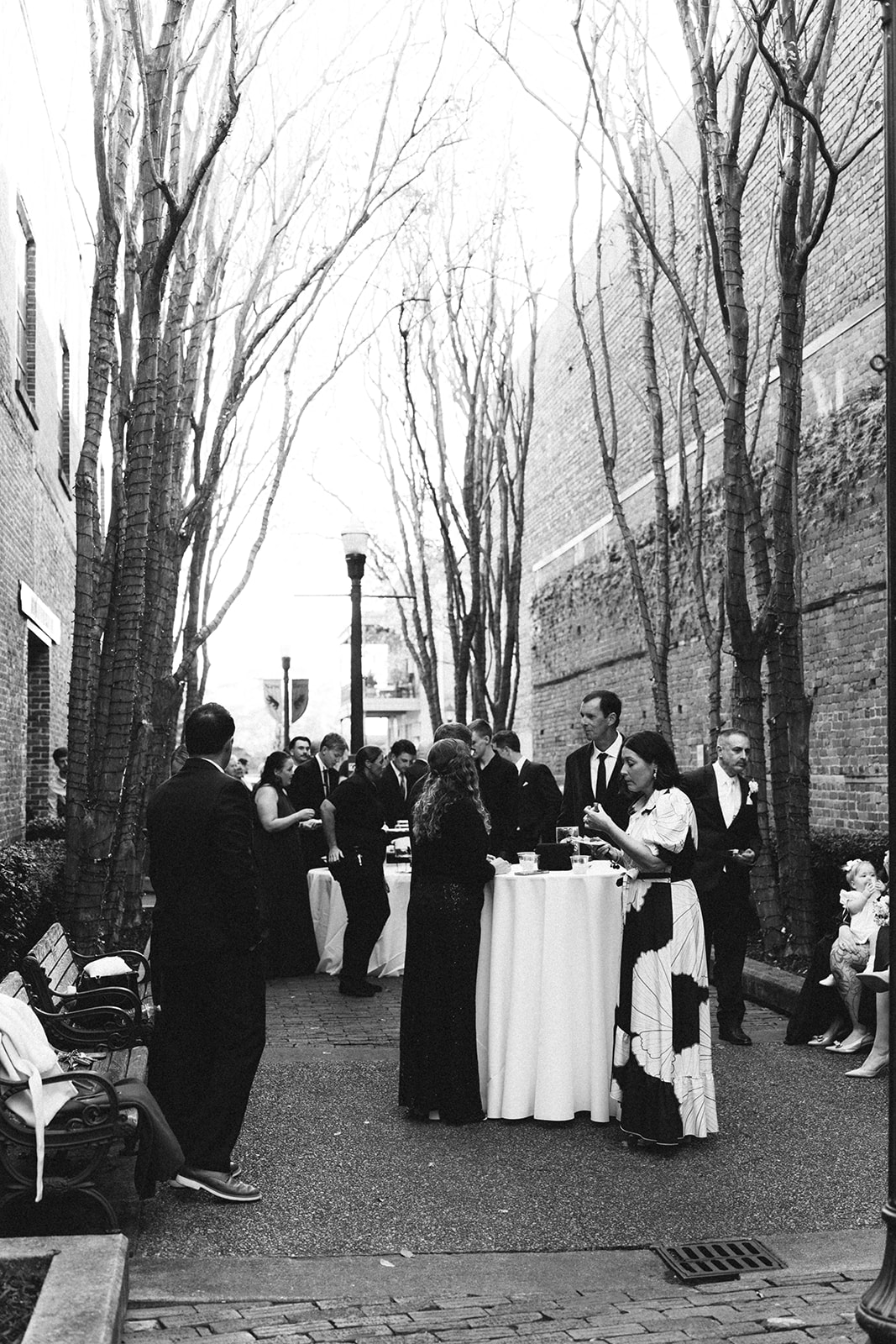 Group of people dressed formally gather around tall cocktail tables in a narrow outdoor alleyway lined with leafless trees and brick buildings.