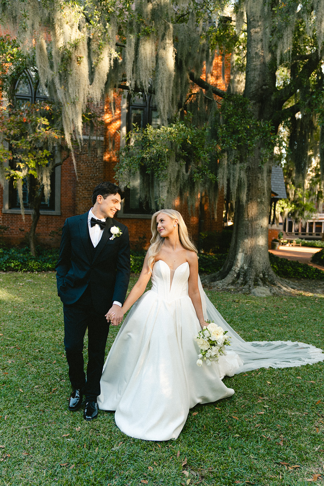 A bride and groom stand outdoors holding hands, smiling at each other, with trees and a building in the background.
