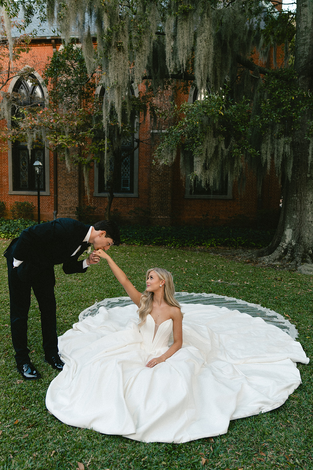 A bride and groom stand outdoors holding hands, smiling at each other, with trees and a building in the background.