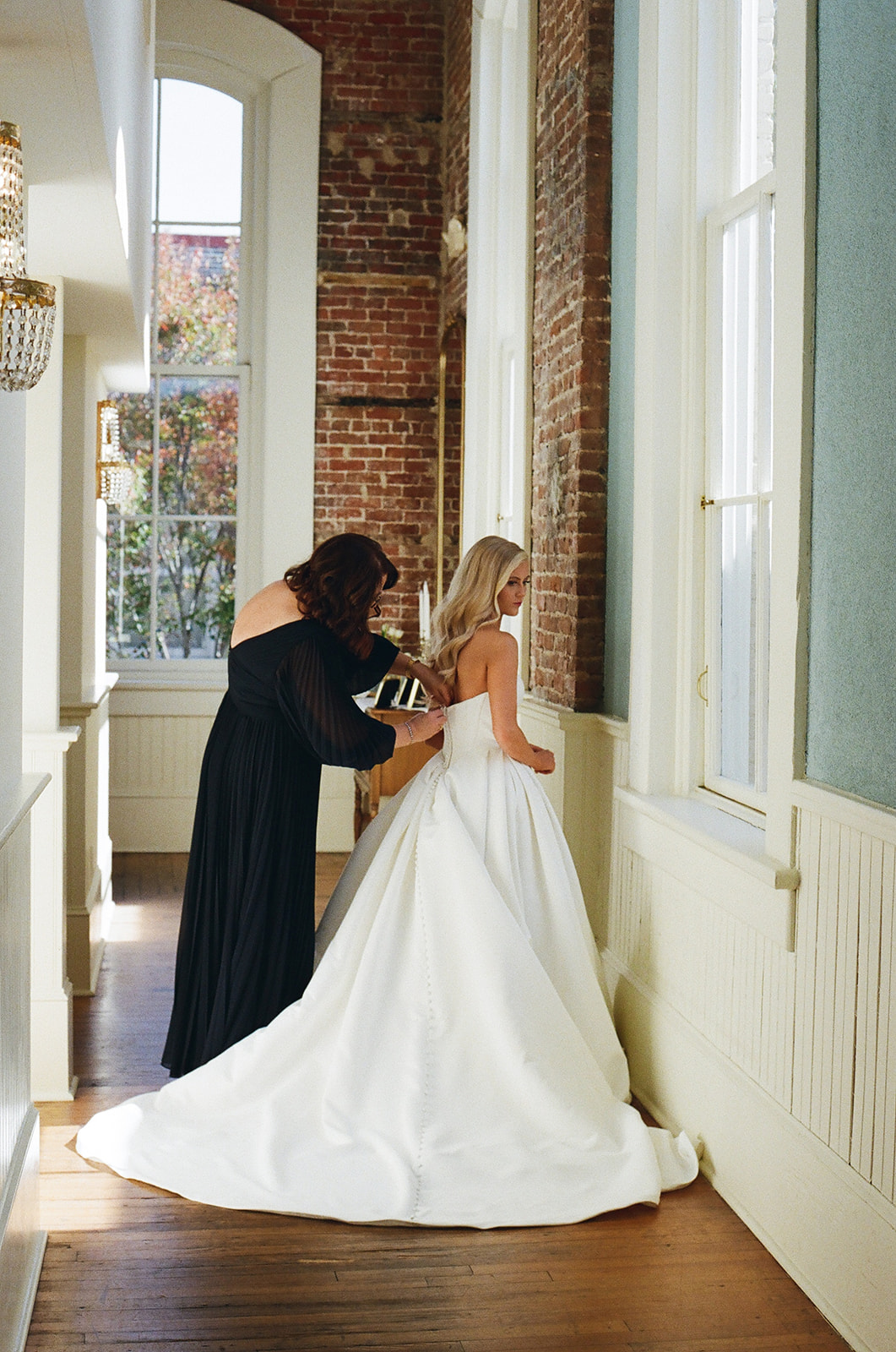 A woman in a white wedding dress stands by a window while another woman adjusts the back of her dress at the Stanly Ballroom a wedding venue in North Carolina